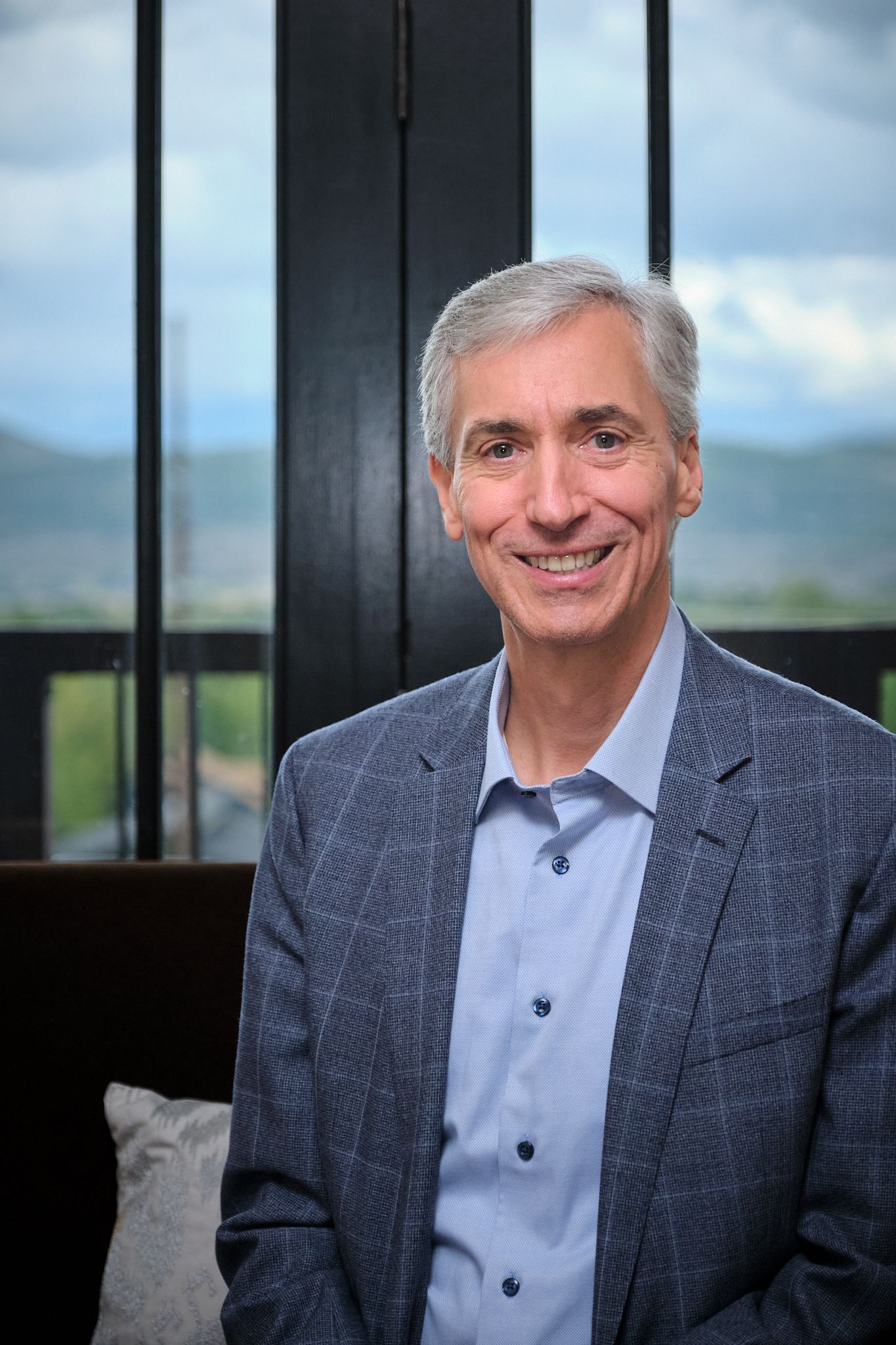 Smiling person with gray hair wearing a light blue shirt and textured blazer, positioned against a windowed landscape.