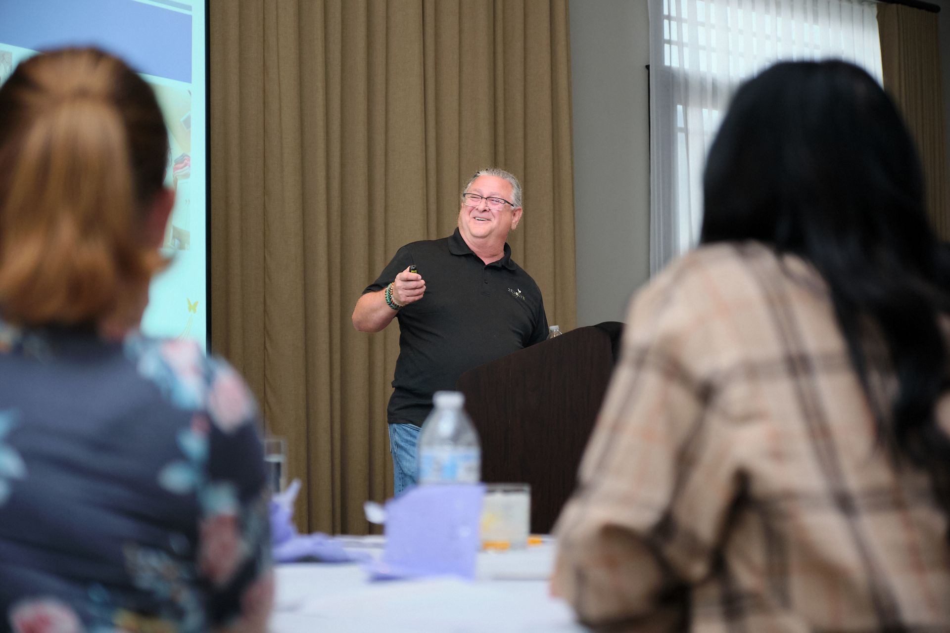 A speaker stands behind a podium, gesturing toward a presentation screen while addressing an audience in a room.