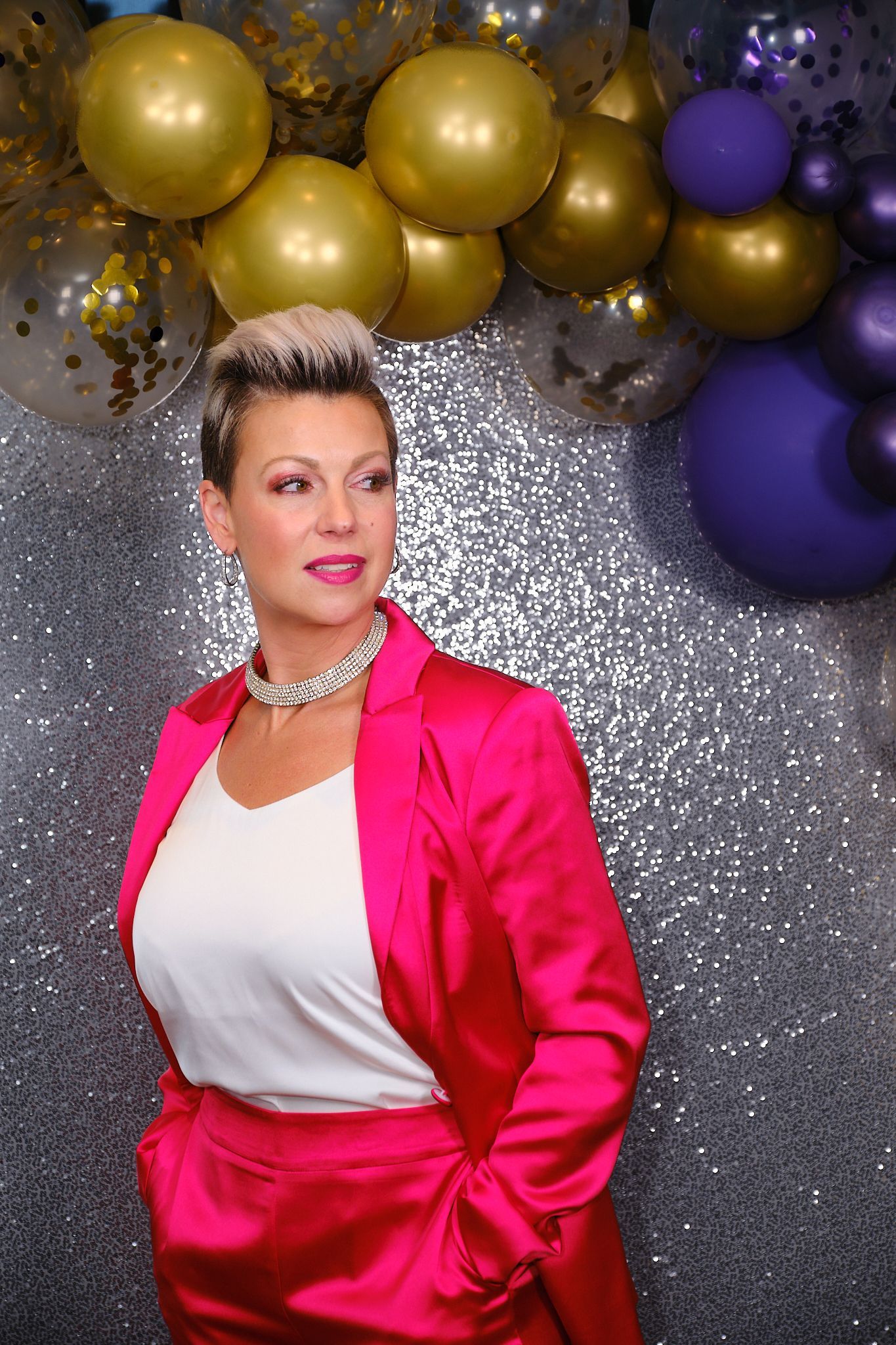 A person with short hair wears a bright pink suit and white top, posing against a silver backdrop with festive balloons.