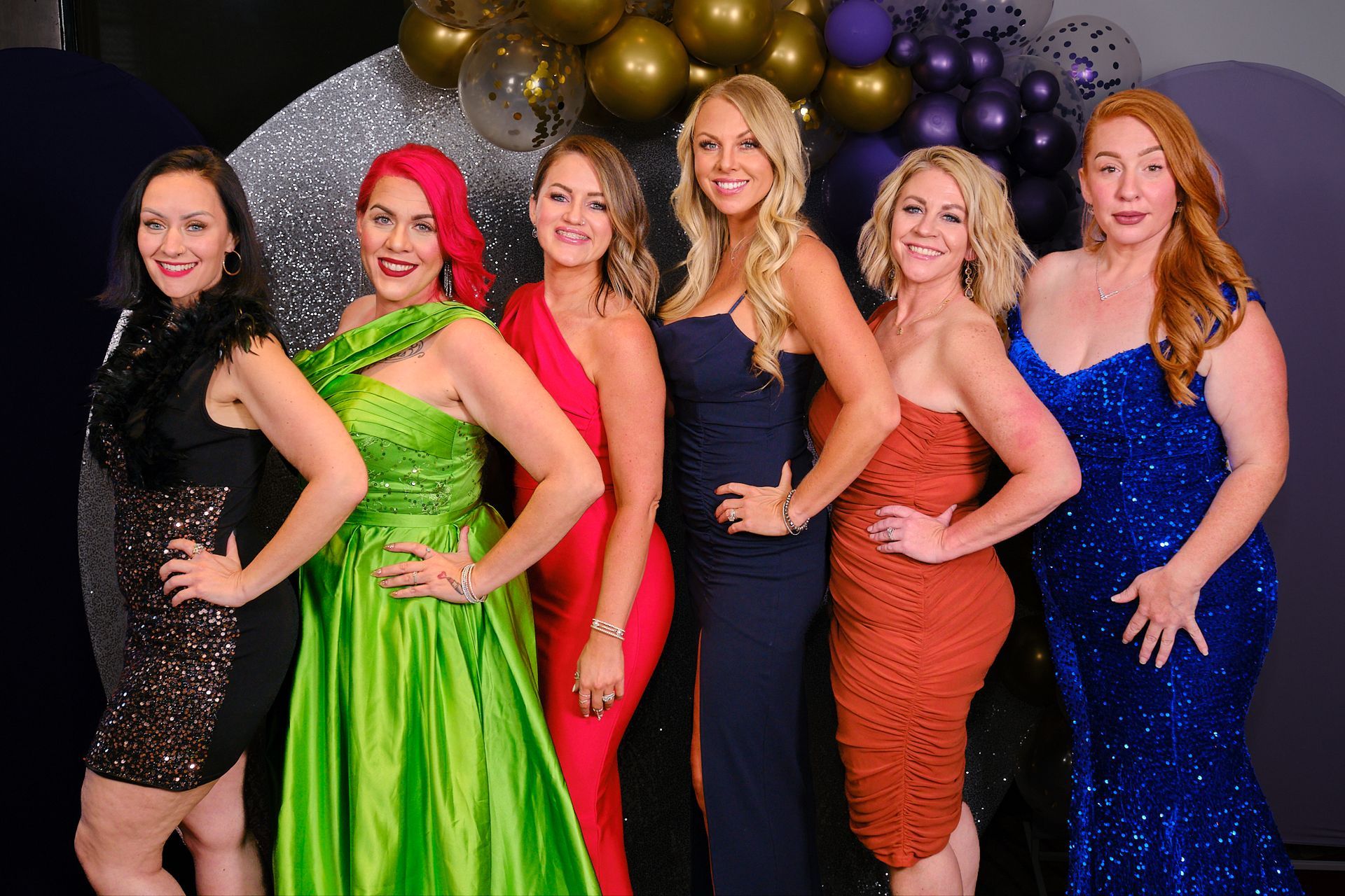 Six women in elegant dresses pose together for a photo in front of a dark backdrop decorated with balloons.