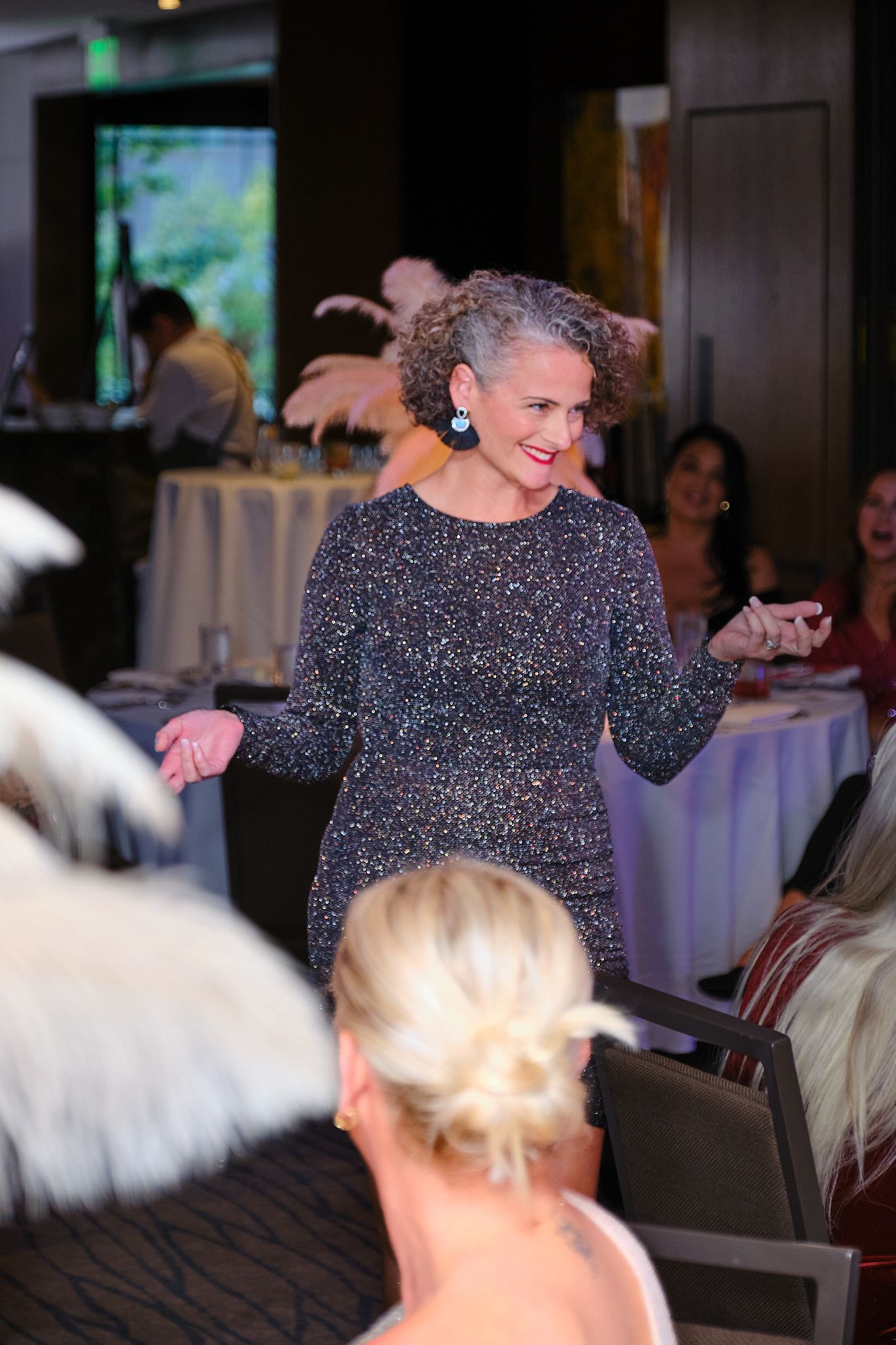 A speaker in a patterned black dress gestures to an audience in a dining room, smiling as she talks.