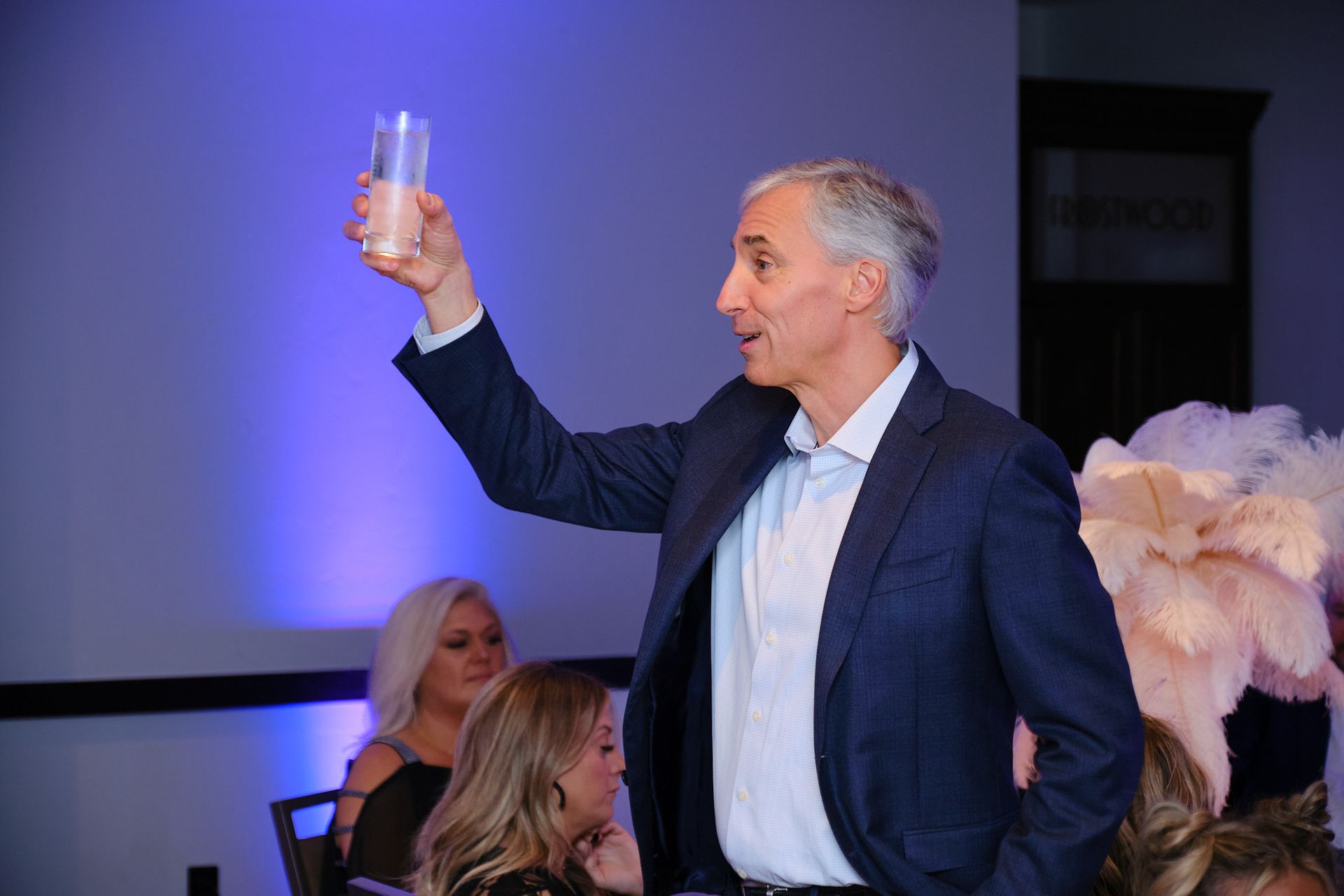A man in a suit raising a glass to offer a toast at an indoor event, with guests seated in the blurred background.