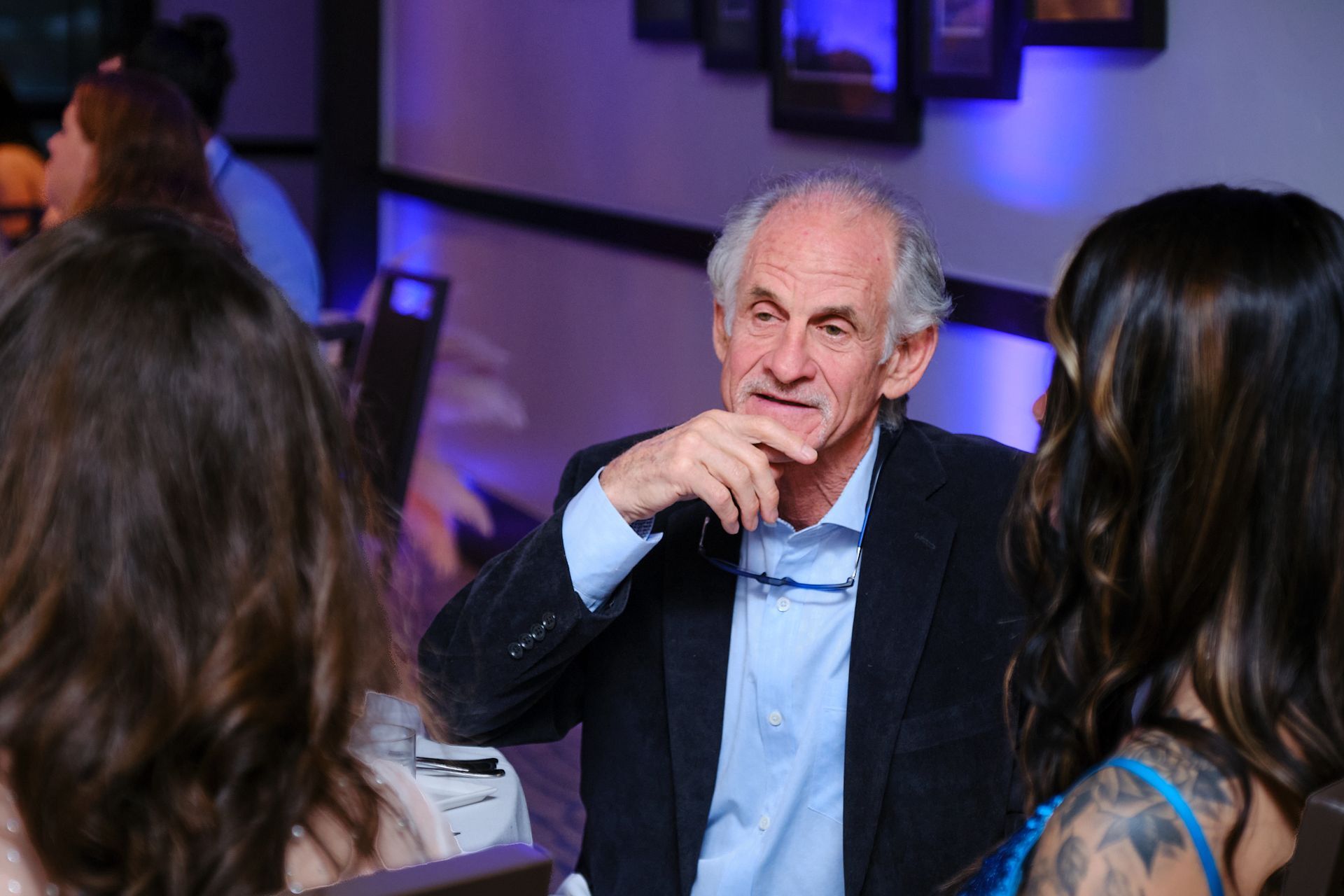 An older person in a suit jacket gestures while speaking to two people at an indoor event with blue lighting.