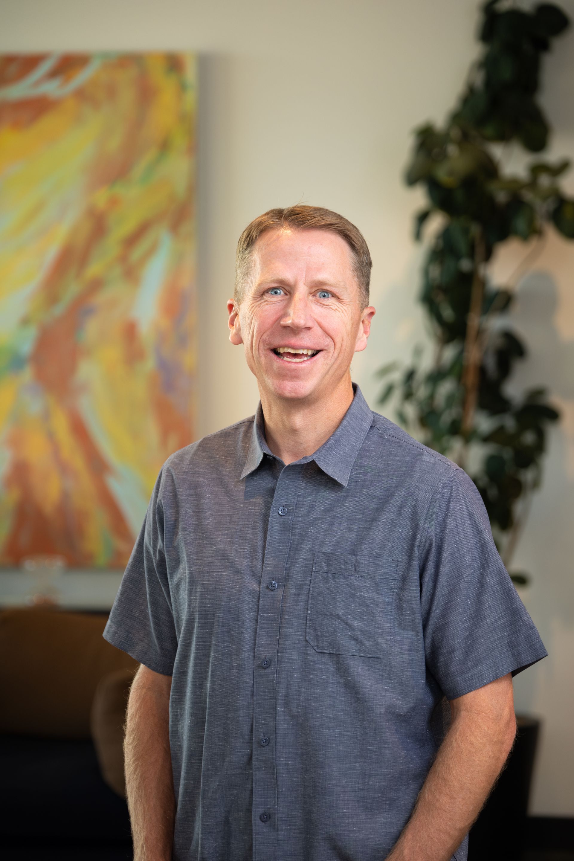 A smiling person in a gray short-sleeved button-down shirt stands in front of a colorful abstract painting and a plant.