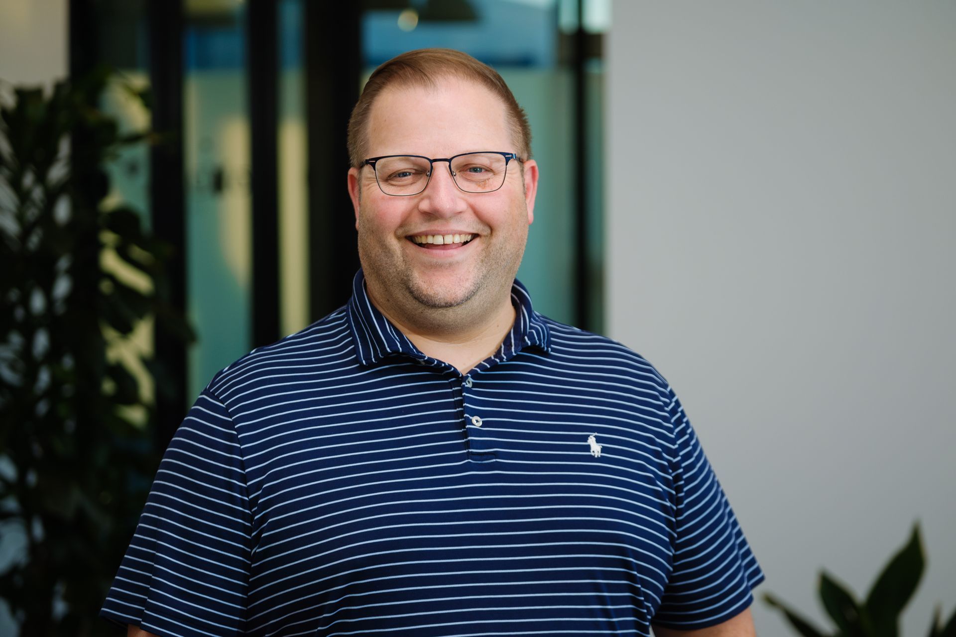 A smiling person wears glasses and a blue-and-white striped polo shirt in an indoor office setting.