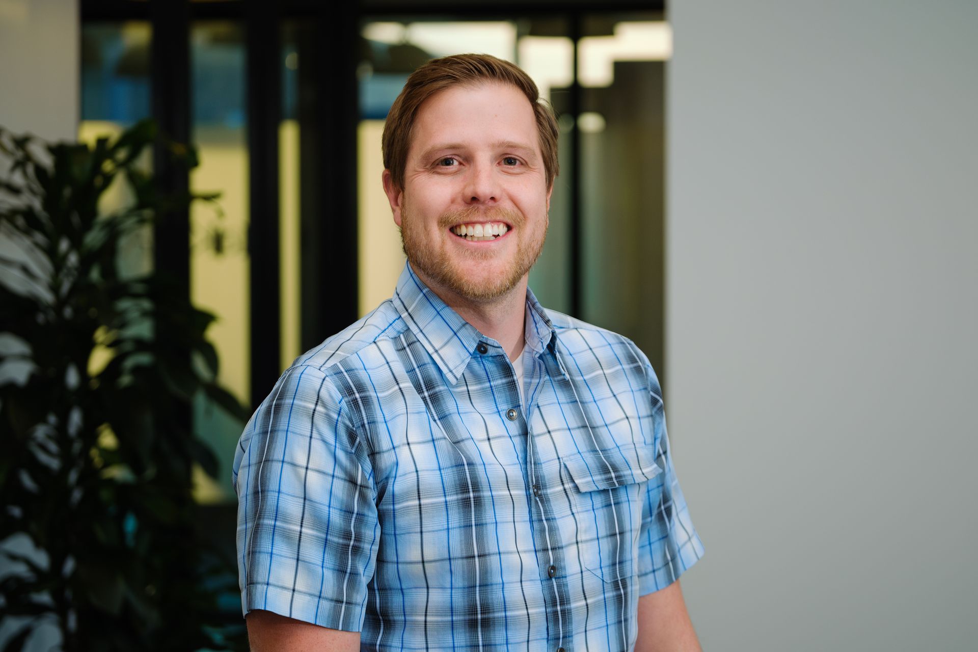 A smiling person in a blue plaid button-down shirt stands in an office setting with a plant in the background.