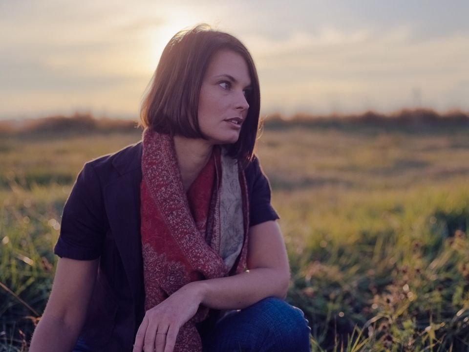 A person with a bob haircut wearing a patterned scarf looks away in a field during sunset.