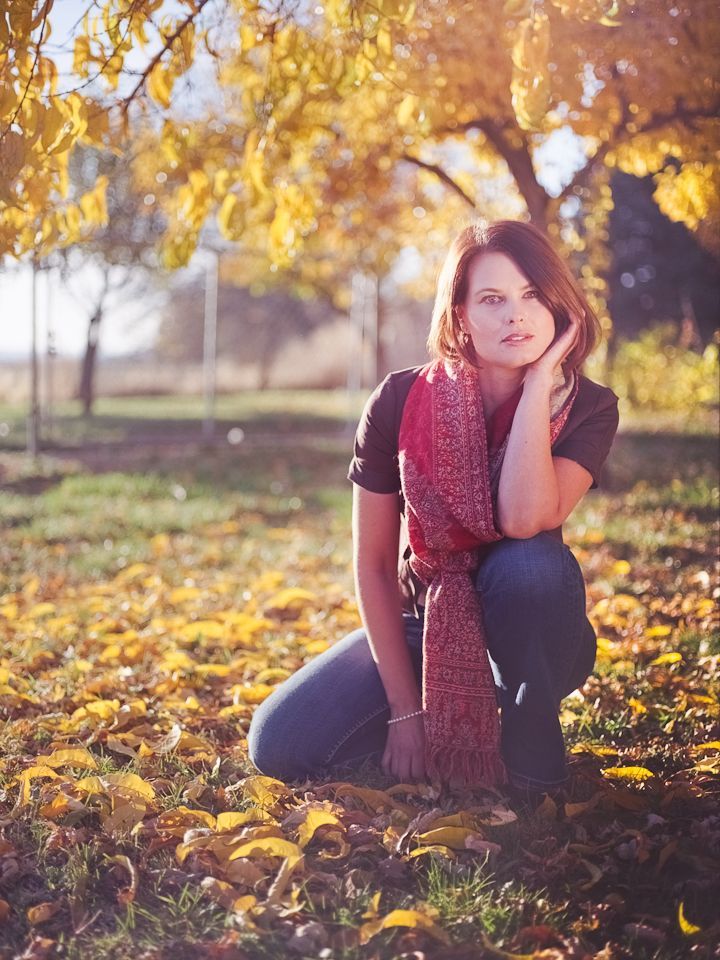 A person wearing a red patterned scarf crouches among golden autumn leaves beneath sunlit trees.