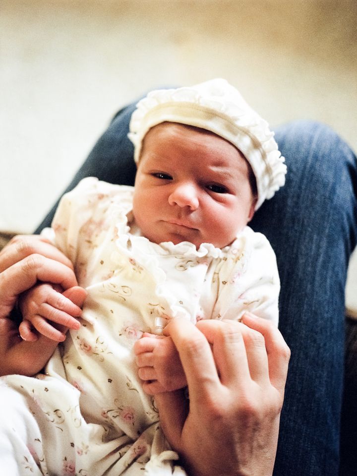 A newborn wearing a white bonnet and patterned outfit being held securely by an adult's hands.