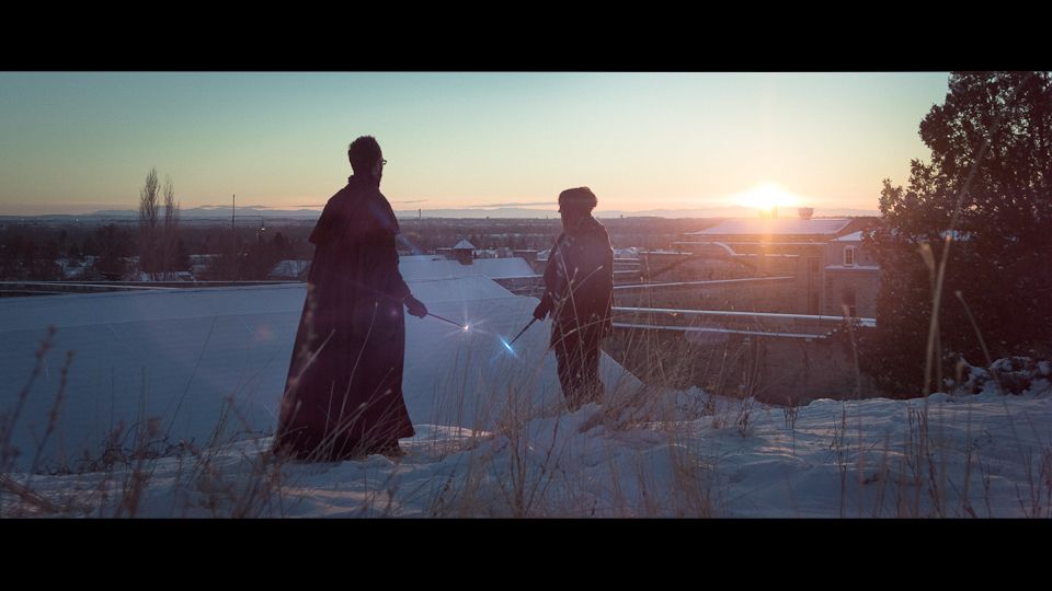 Two figures in dark, flowing robes stand on a snowy hill at sunset, pointing glowing wands toward each other.