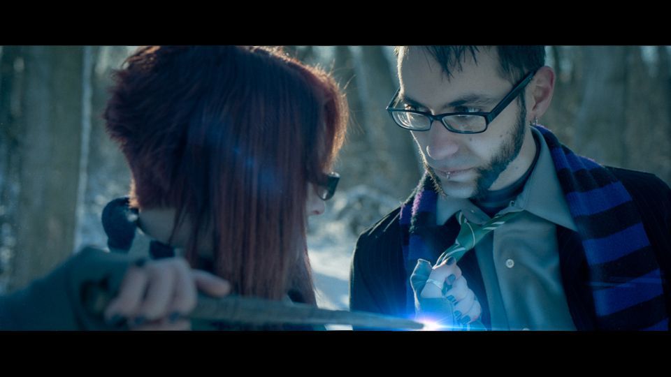 A person holds a glowing blade toward another person in a forest, both wearing glasses and scarves in a cinematic shot.
