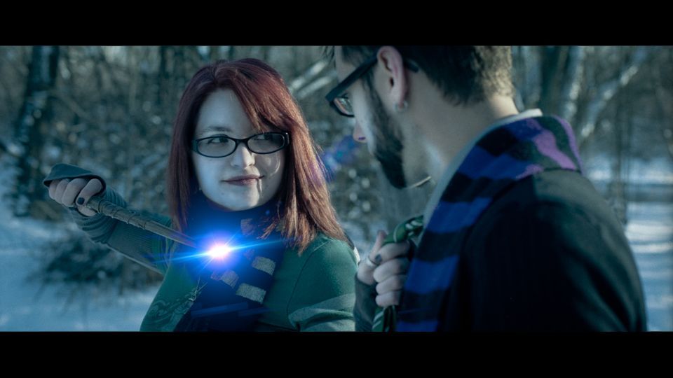 A person holds a glowing item toward another in a snowy, wooded area, wearing a green coat and dark-rimmed glasses.