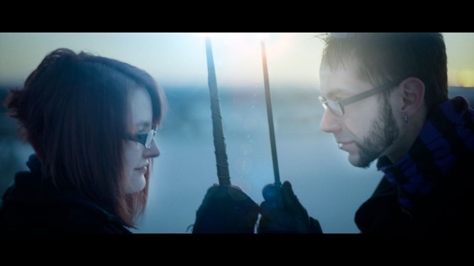 Two people face each other in a landscape, holding up wands in a backlight, wintry setting.