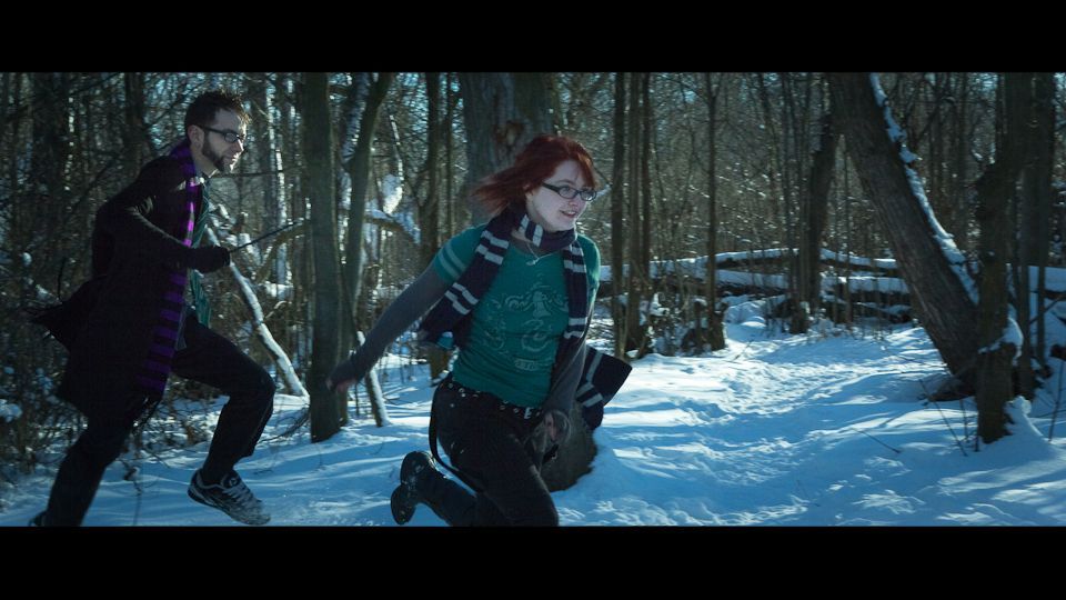 Two people wearing scarves run through a snow-covered, sunlit forest.