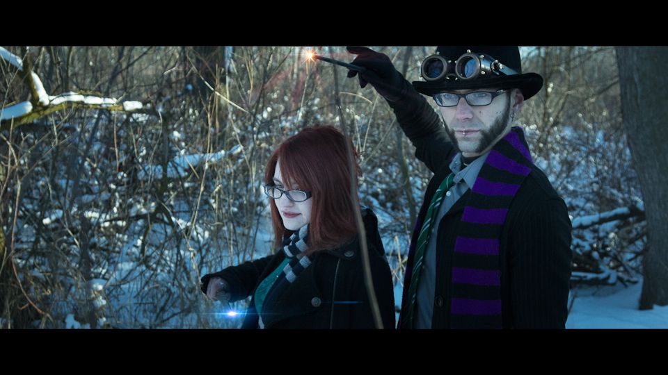 Two people in wizard attire cast glowing spells with wands in a snowy, wooded area.