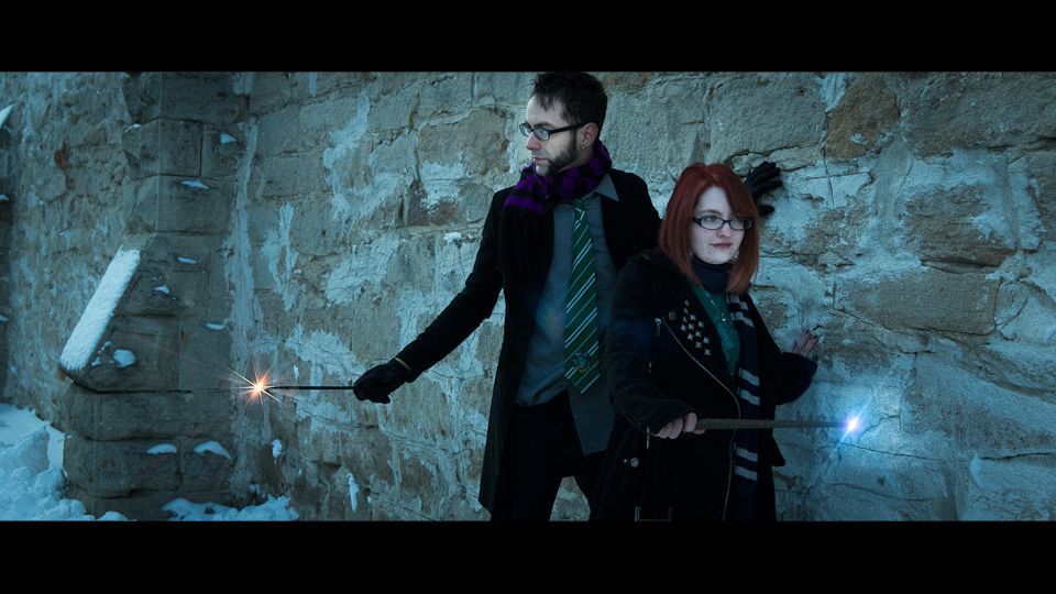 Two people in coats and scarves stand against a stone wall, holding glowing wands in a snowy, wintry setting.