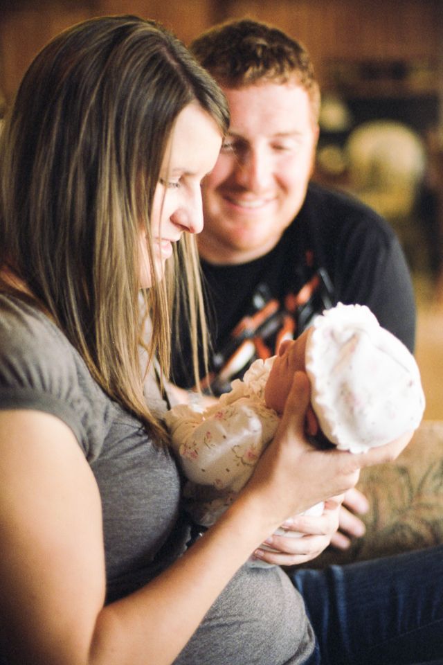 A parent cradling a newborn wearing a white cap while another parent watches with a smile.