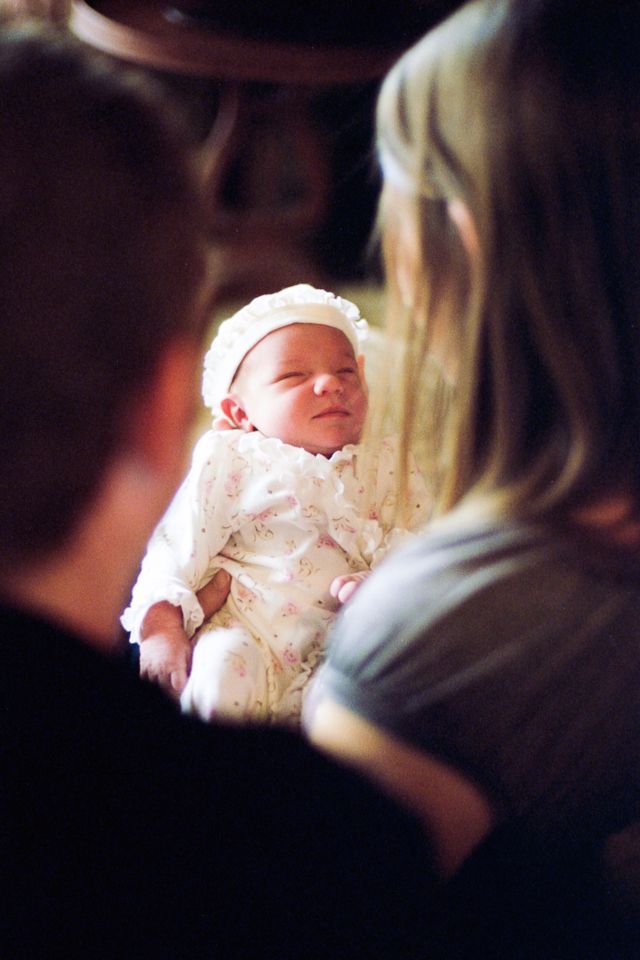 A baby wearing a bonnet and floral outfit is held by two adults, seen from behind.