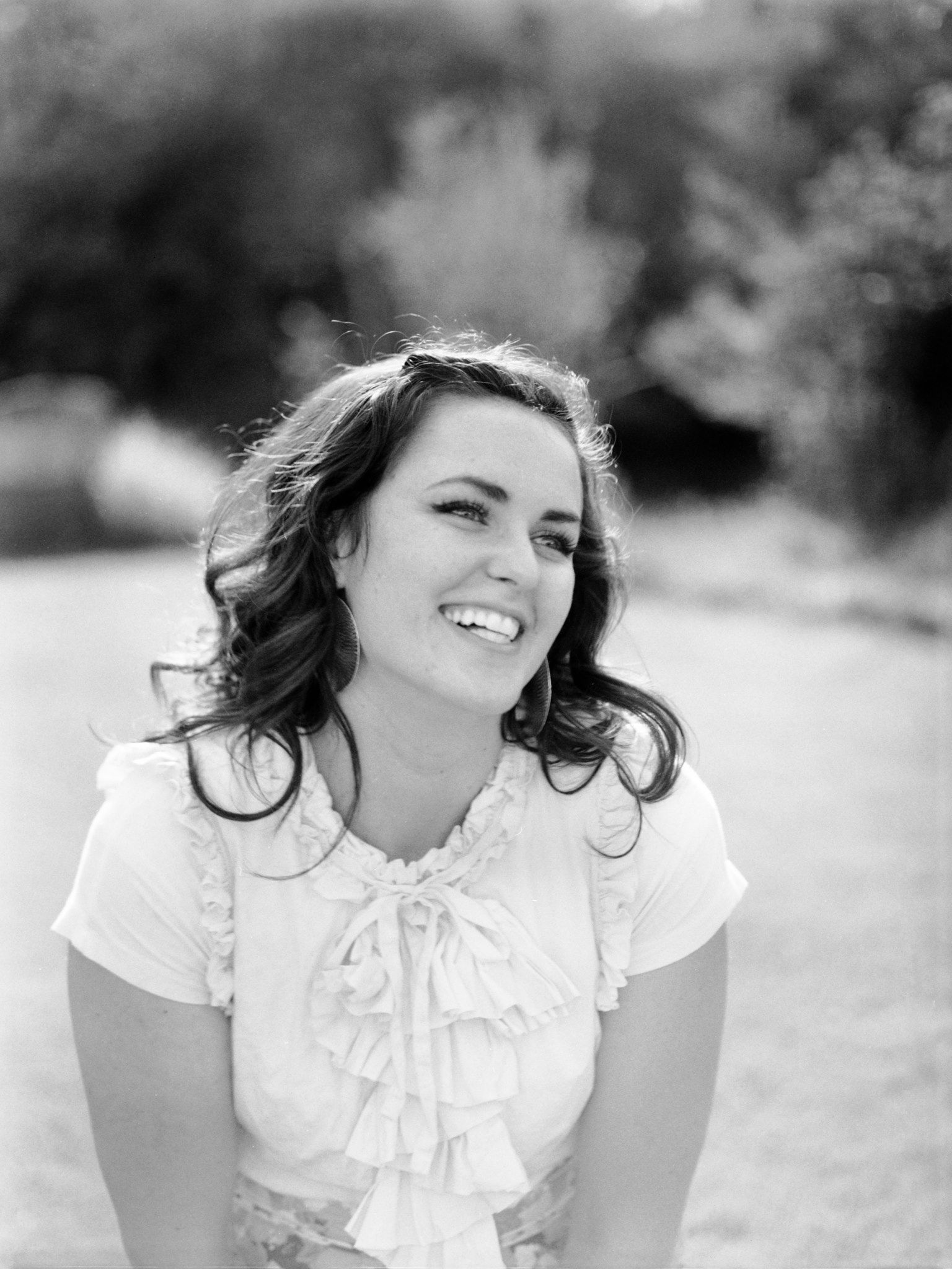 Black and white portrait of a smiling person with wavy hair, wearing a ruffled blouse outdoors in a soft-focus setting.