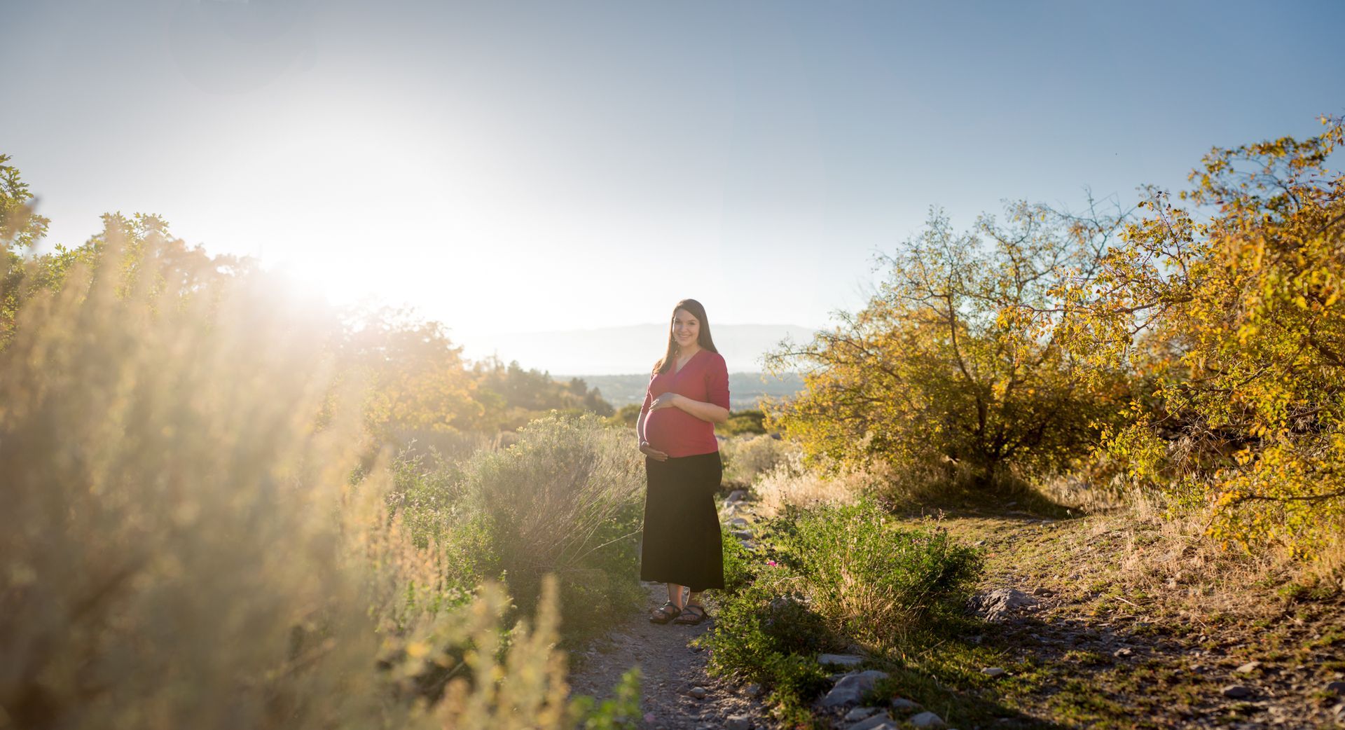 A pregnant person in a red top and black skirt stands on a sunlit path surrounded by bushes in an open field.