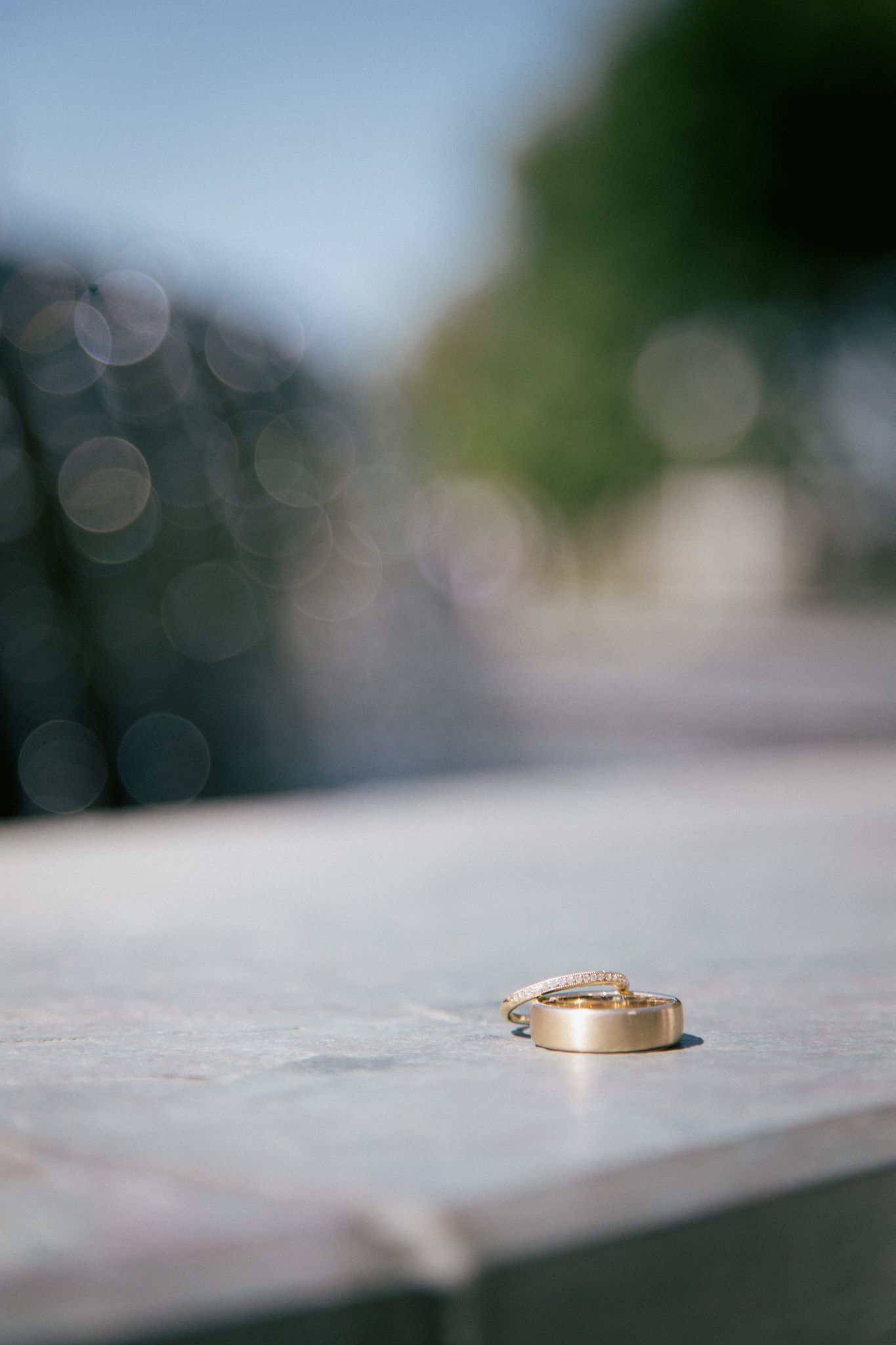 Two gold wedding rings rest on a smooth, light-colored surface with a blurred outdoor background.