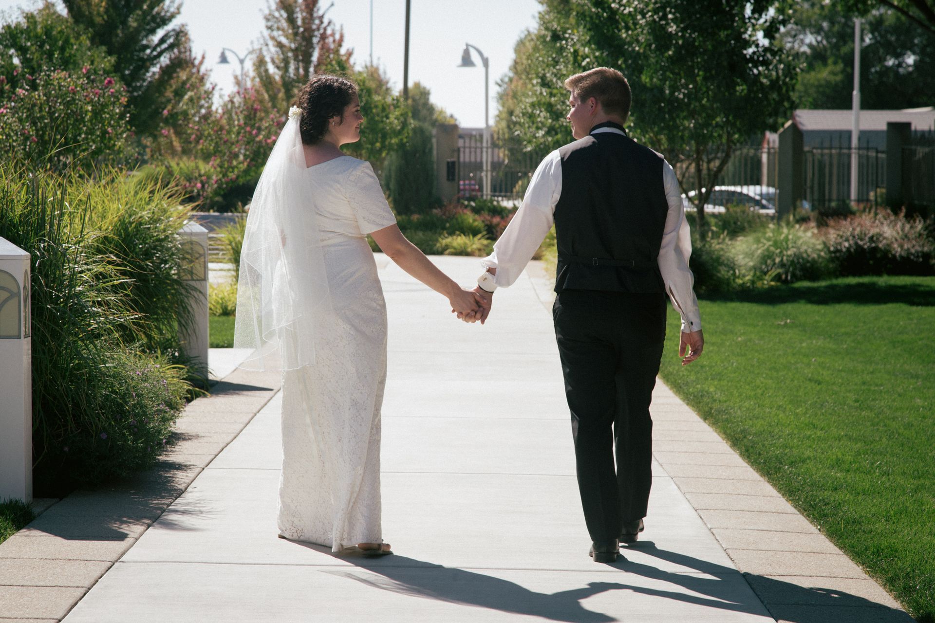 A couple in wedding attire walks hand-in-hand along a sunny outdoor path, looking back toward one another.