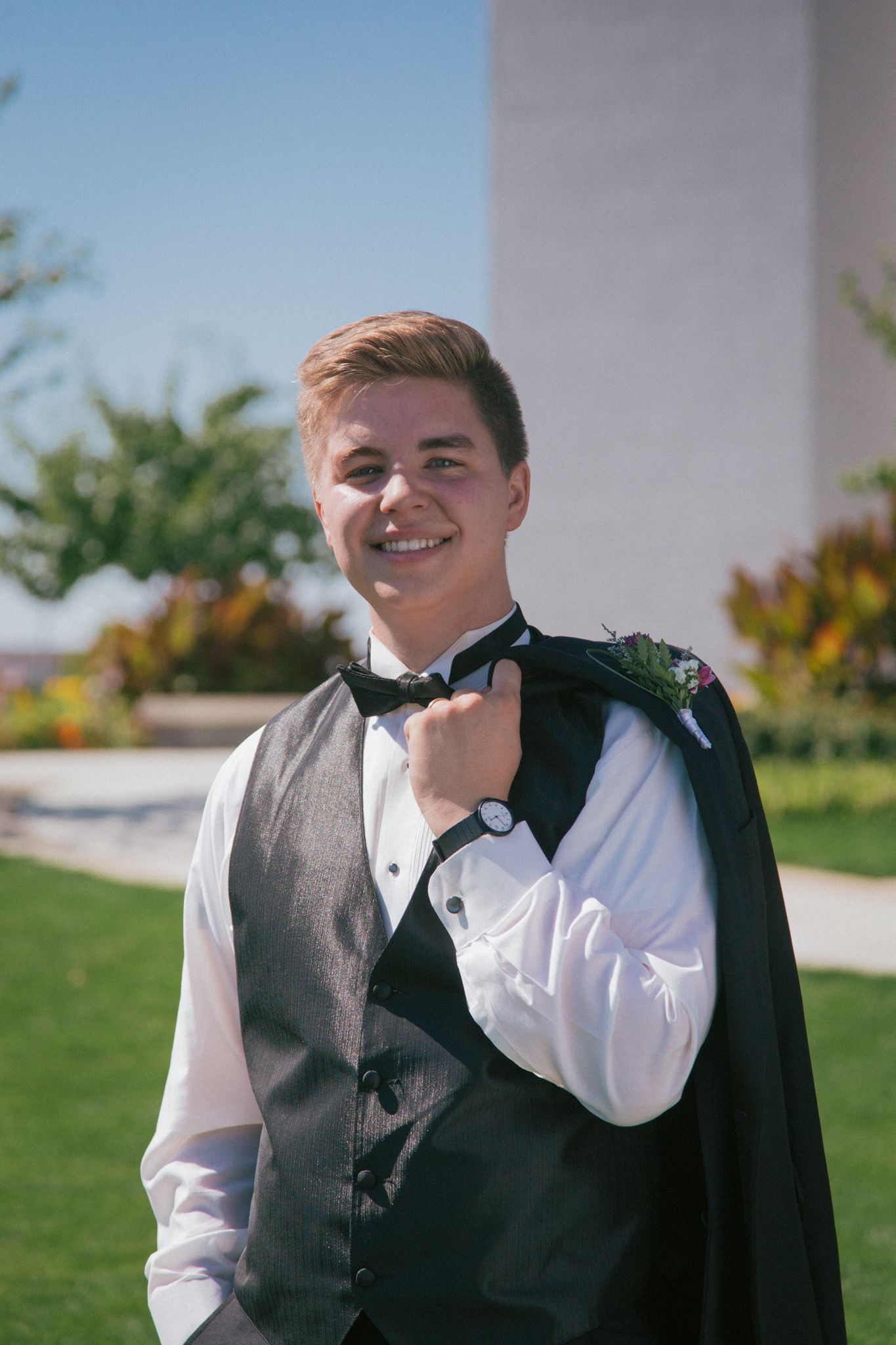 A person wearing a tuxedo vest, white dress shirt, and bowtie, smiling while holding a suit jacket over their shoulder.