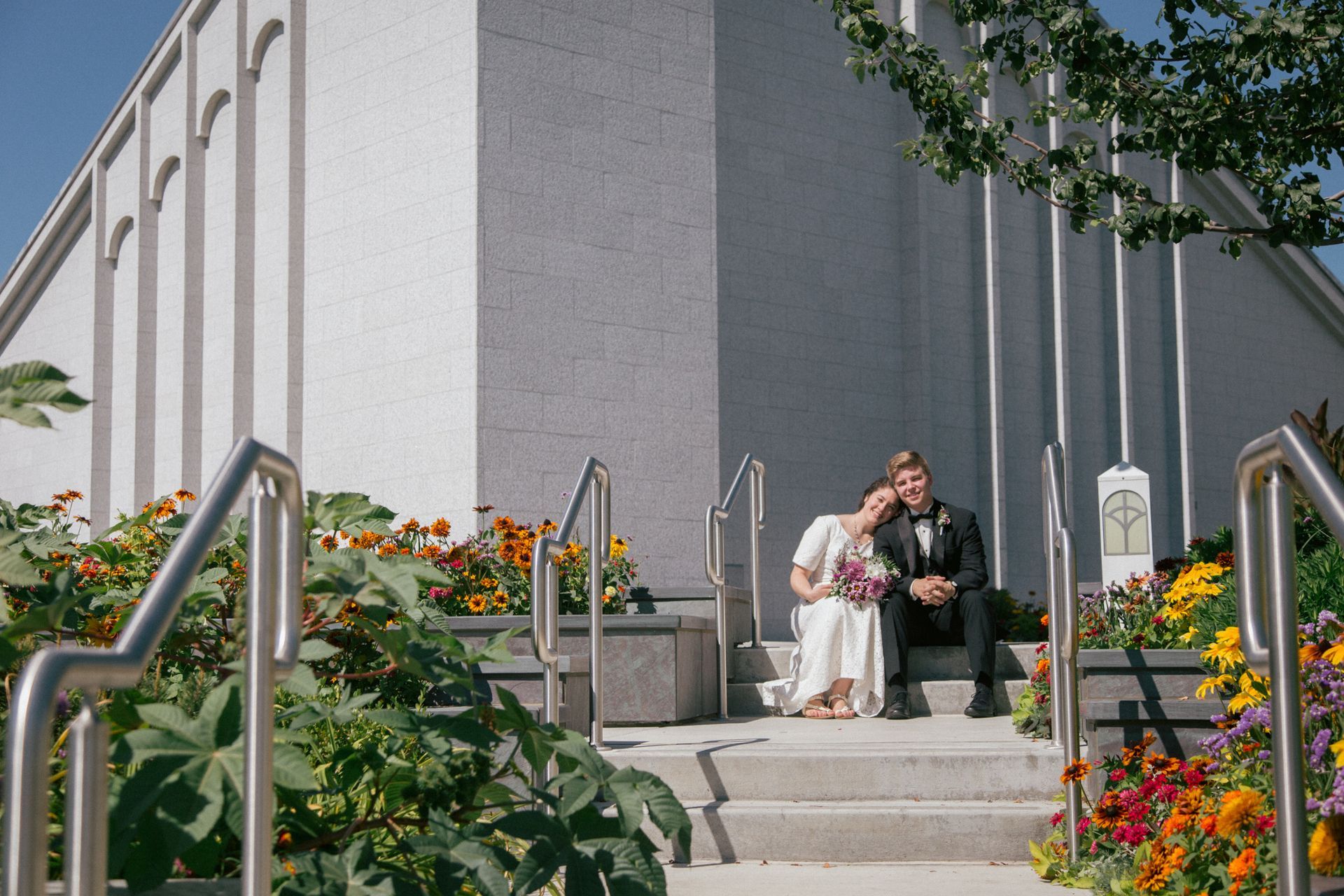 A couple sitting on steps of a modern building surrounded by colorful flowers on a sunny day.