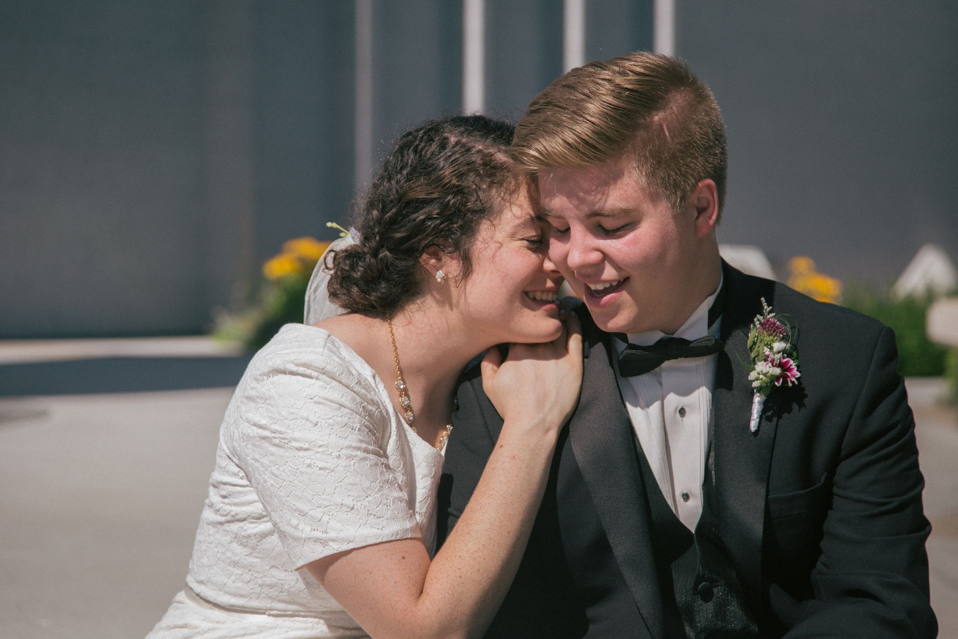 A person in a white lace dress leans their head on the shoulder of another person in a black tuxedo with a boutonniere.