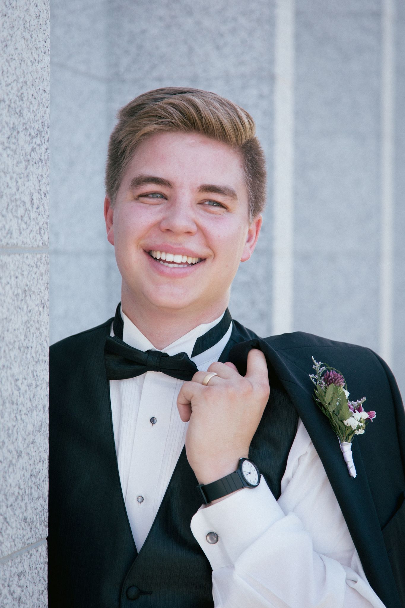 A smiling person in a tuxedo and bowtie holds a black jacket over their shoulder while standing against a stone wall.