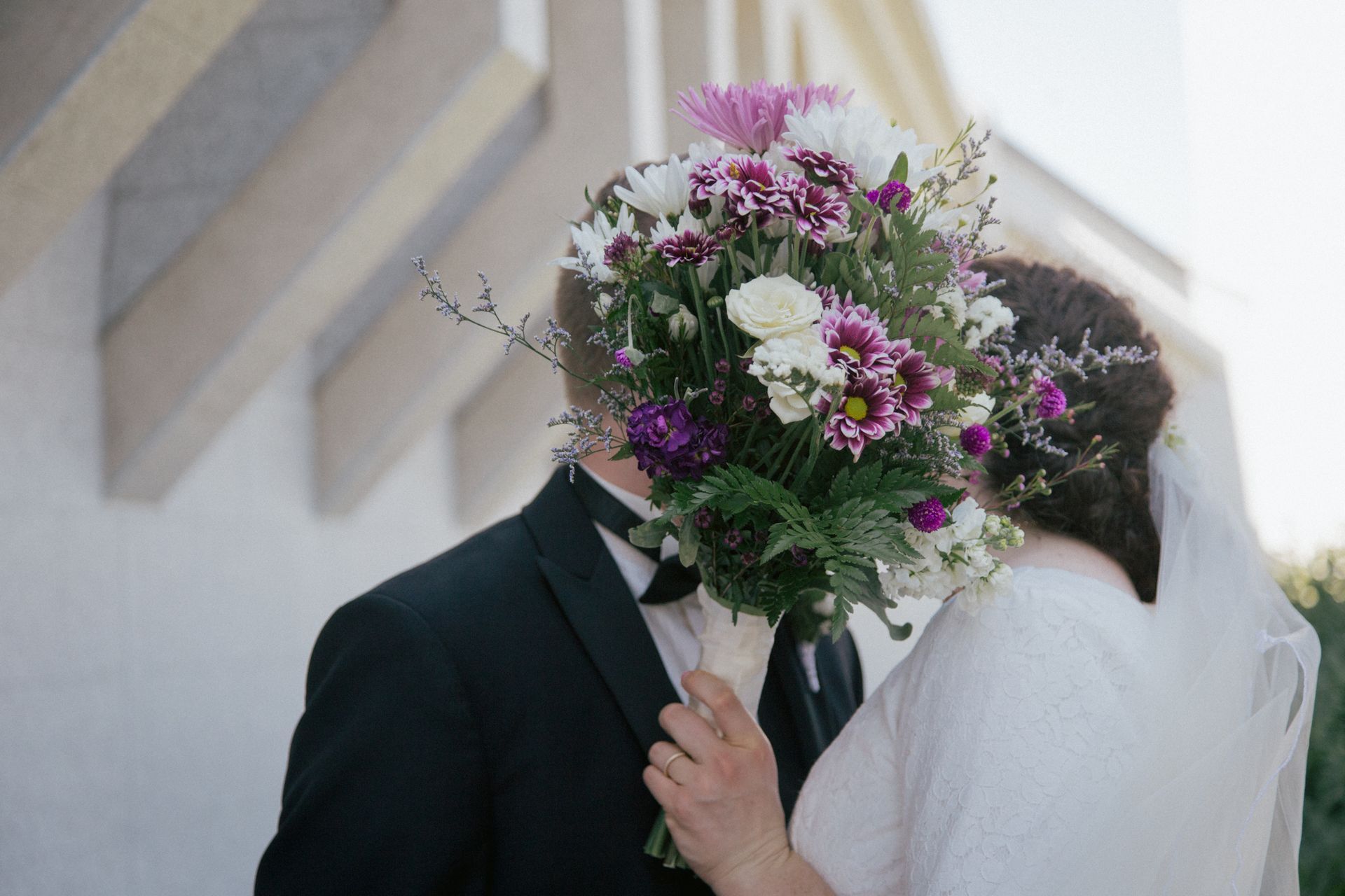 A bride and groom kiss behind a large bouquet of purple and white flowers.