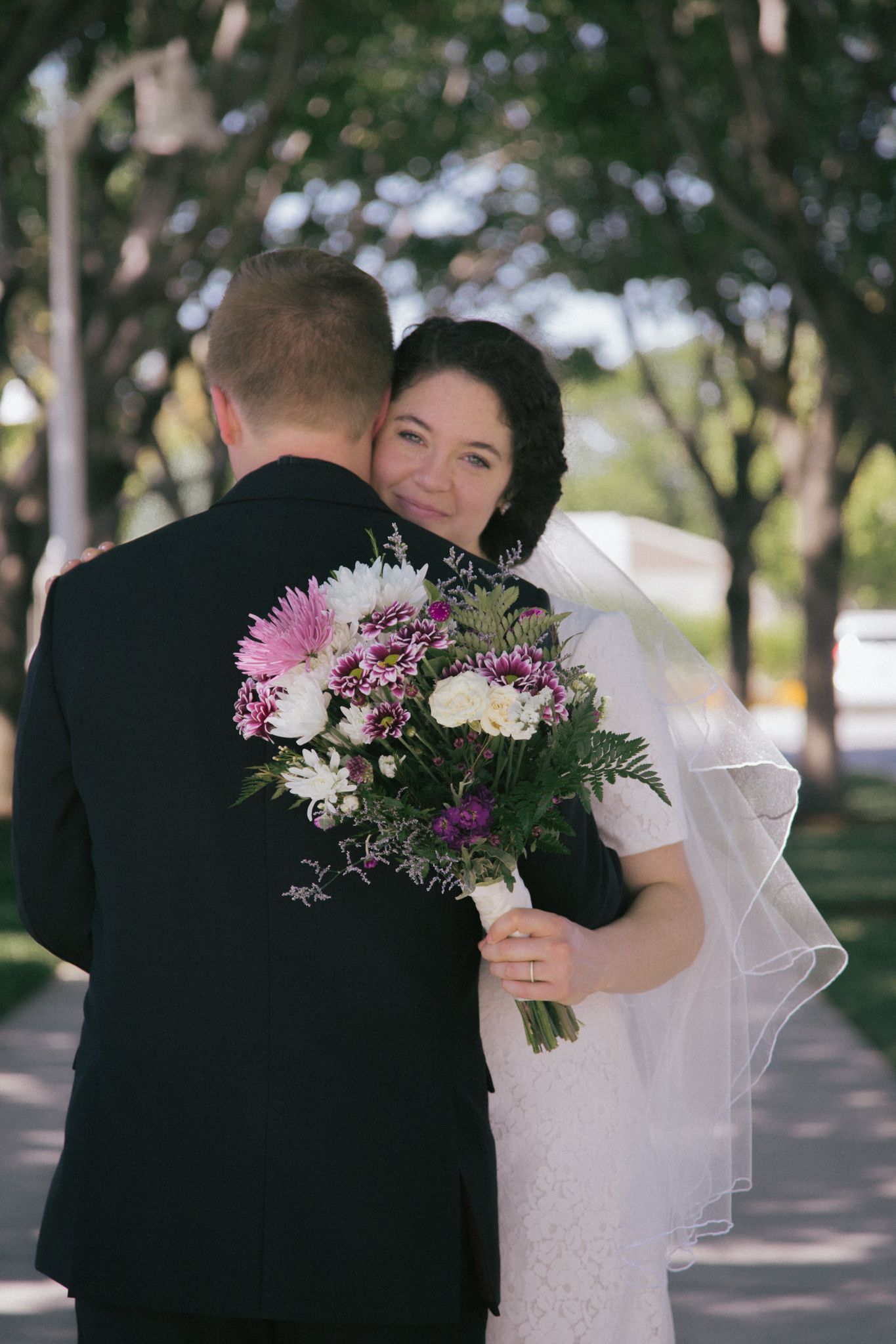 A person in a wedding gown holding a floral bouquet embraces another person in a black suit outdoors.