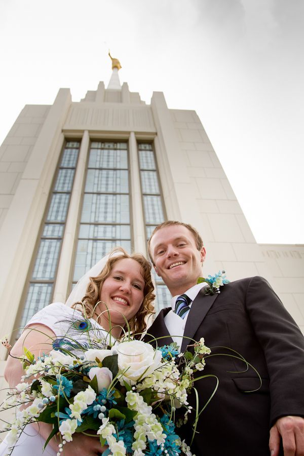 A smiling couple in wedding attire holds a white and blue floral bouquet in front of a tall, stone temple.