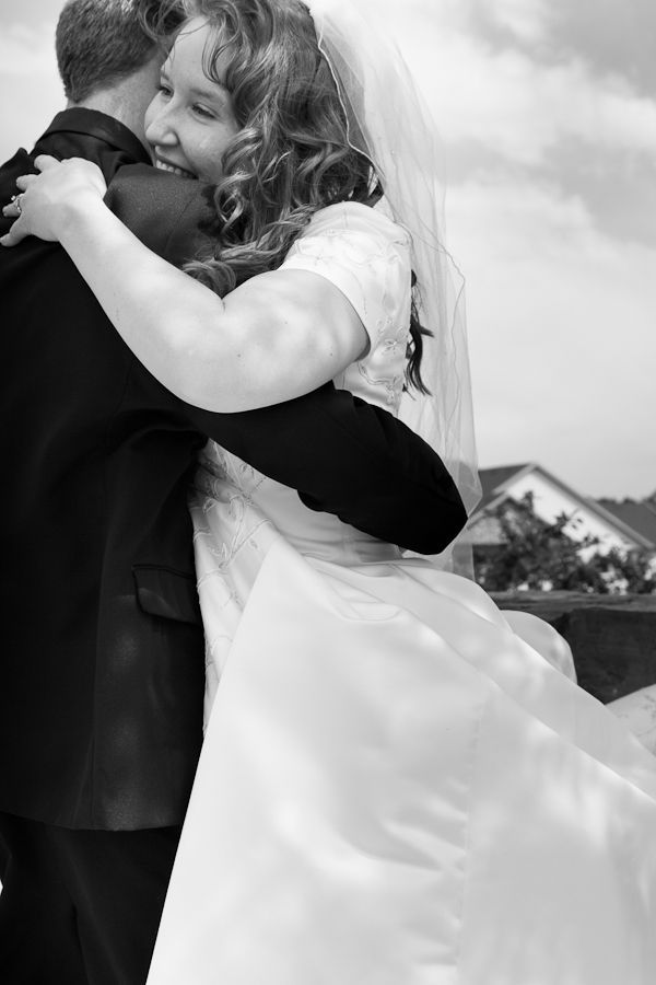 A black-and-white photo of a bride in a gown and veil hugging a person wearing a dark suit outdoors.