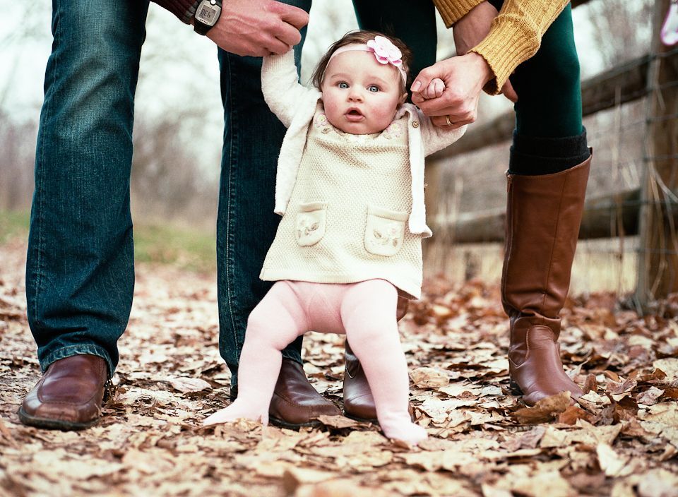 Parents hold their young child’s hands while walking through a pile of dry autumn leaves.