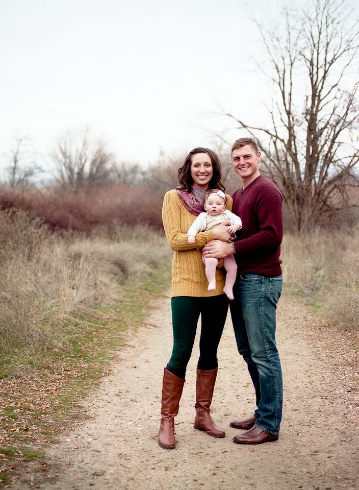 A family of three stands on a dirt path in a park, smiling at the camera, with bare trees in the background.