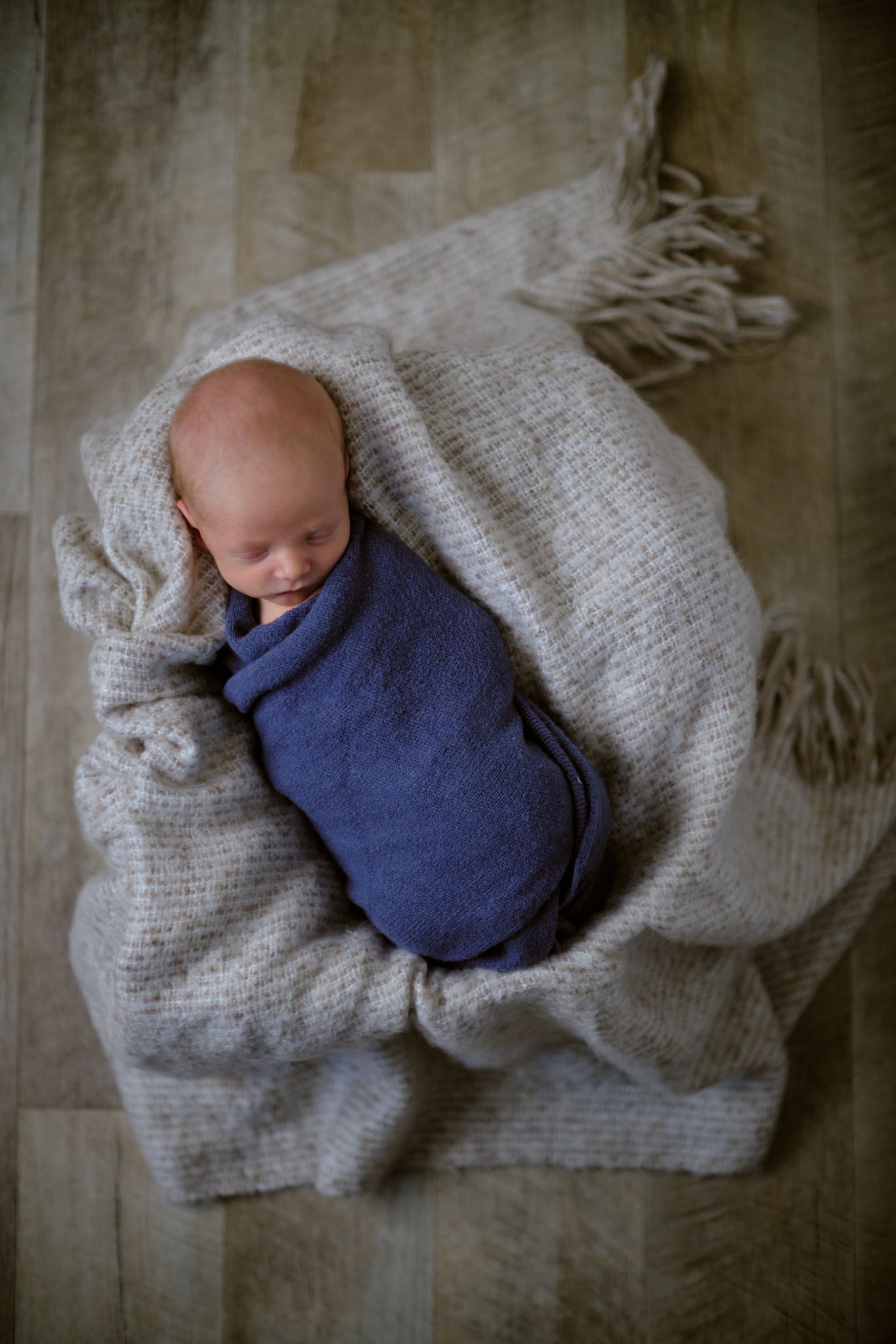 A newborn is wrapped in a blue swaddle, resting on a textured, light-colored fringed blanket on a wooden floor.