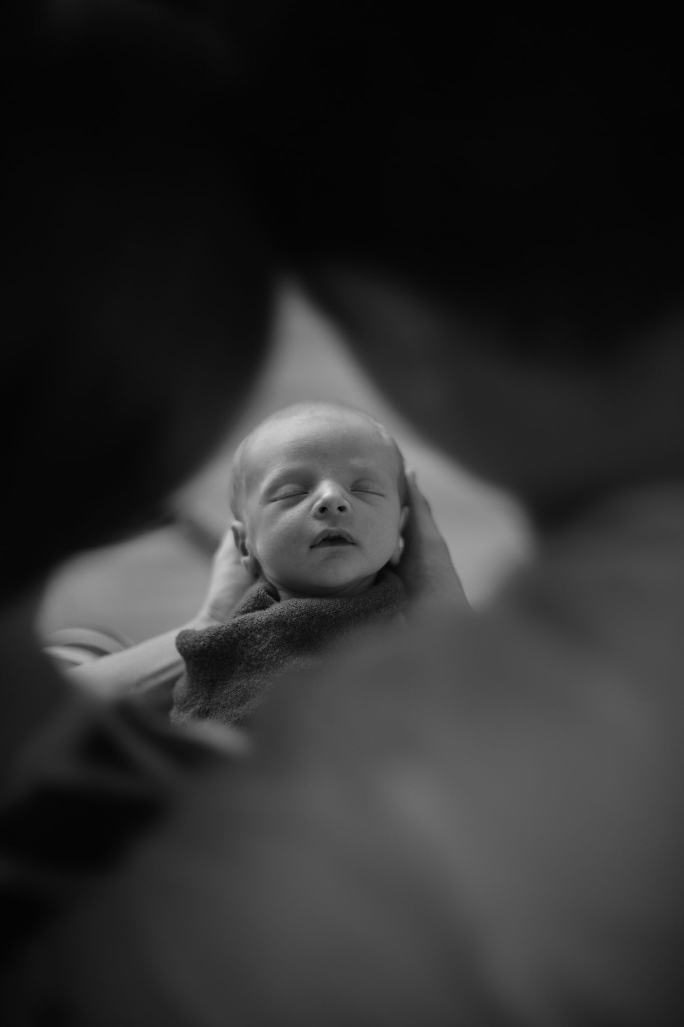 A black-and-white close-up of a newborn held in cupped hands, framed by blurred silhouettes of two people.