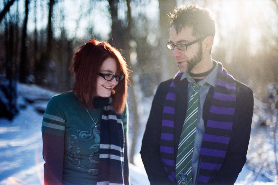 A man and woman wearing striped scarves and glasses stand outdoors in a snowy, sunlit wooded area, looking at each other.