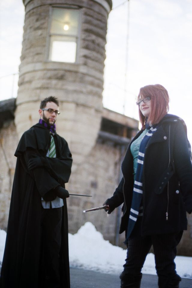 Two people dressed in Harry Potter-inspired wizard costumes stand outdoors holding wands before a stone tower in winter.