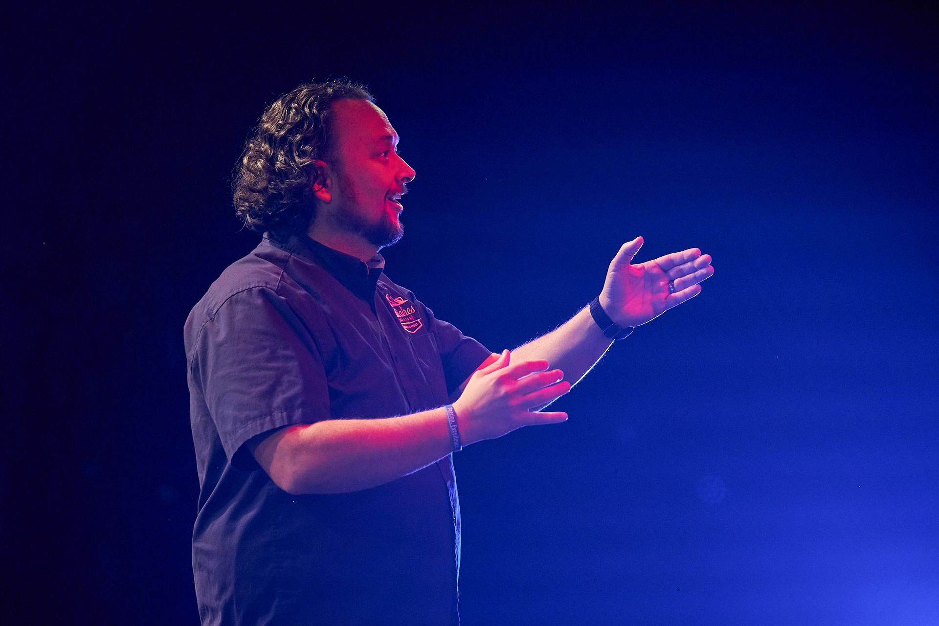 A person stands on a stage, gesturing with both hands while speaking, illuminated by a deep blue light.