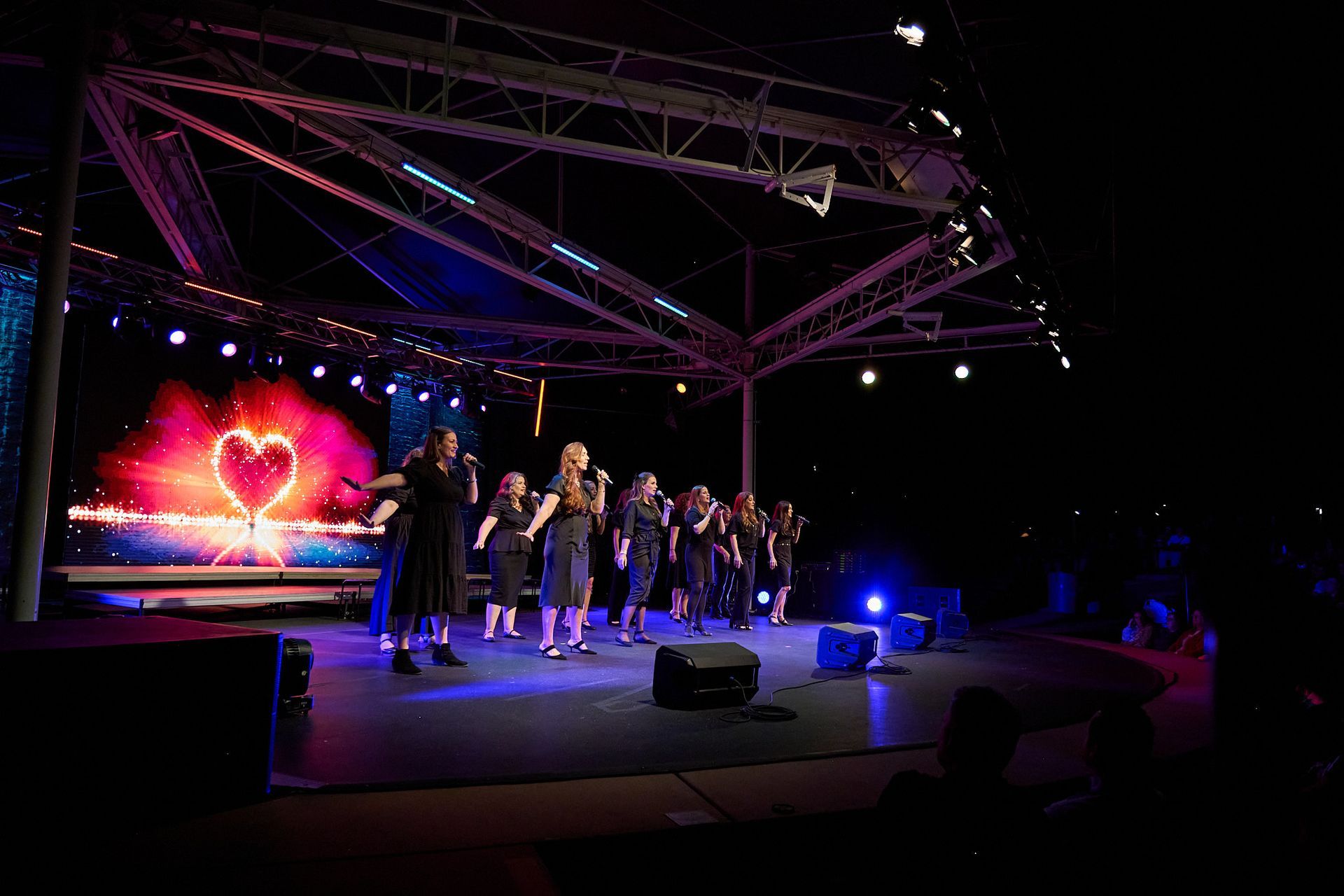 A group of people singing in unison on an outdoor stage with a large heart graphic displayed on a screen behind them.
