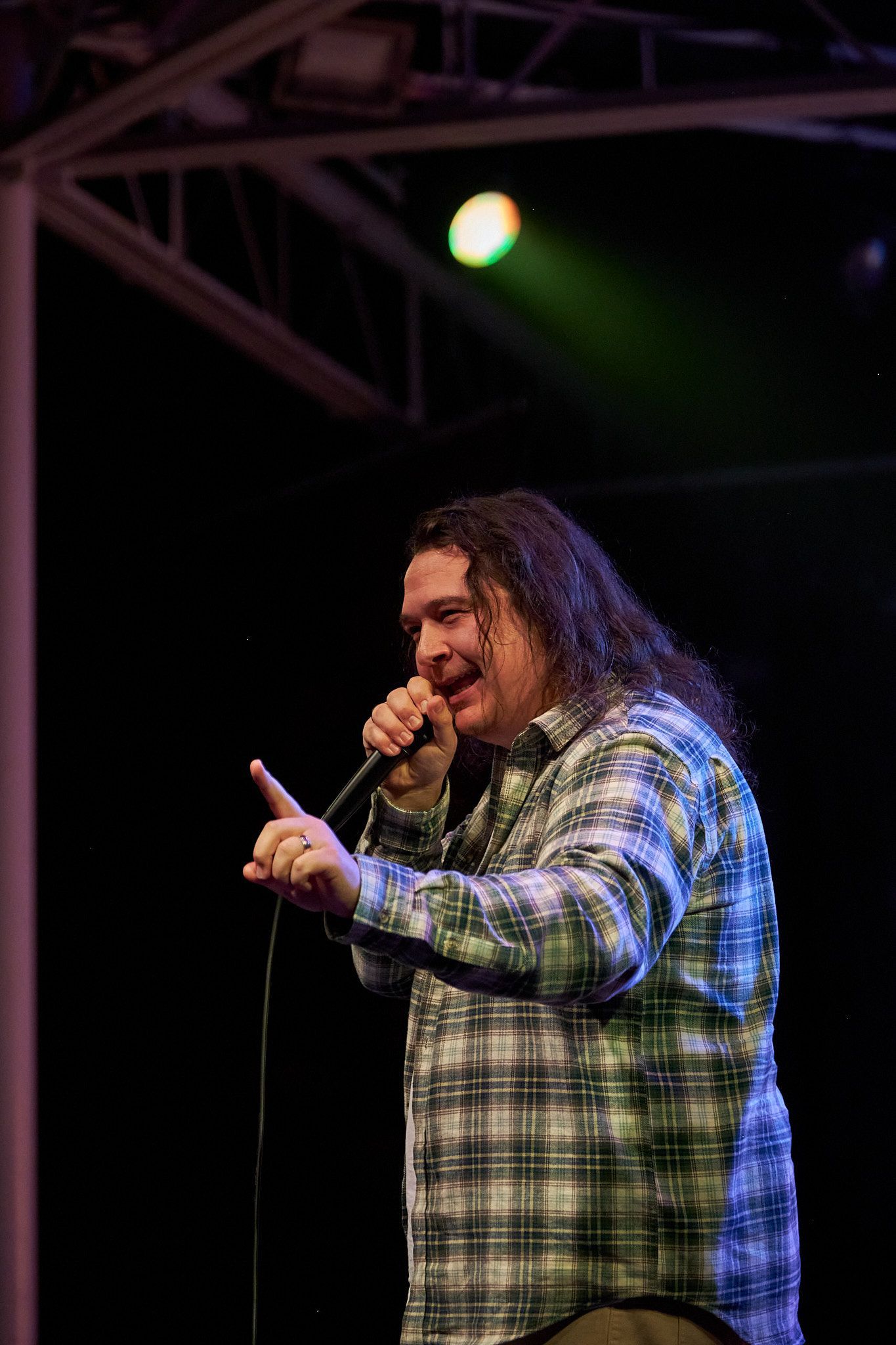 A person with long hair wears a green and blue plaid shirt, speaking into a microphone on stage under a spotlight.