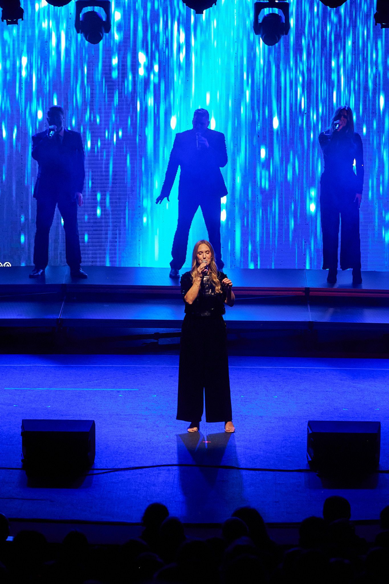 A soloist sings on a blue-lit stage in front of three silhouettes, with two black speakers in the foreground.