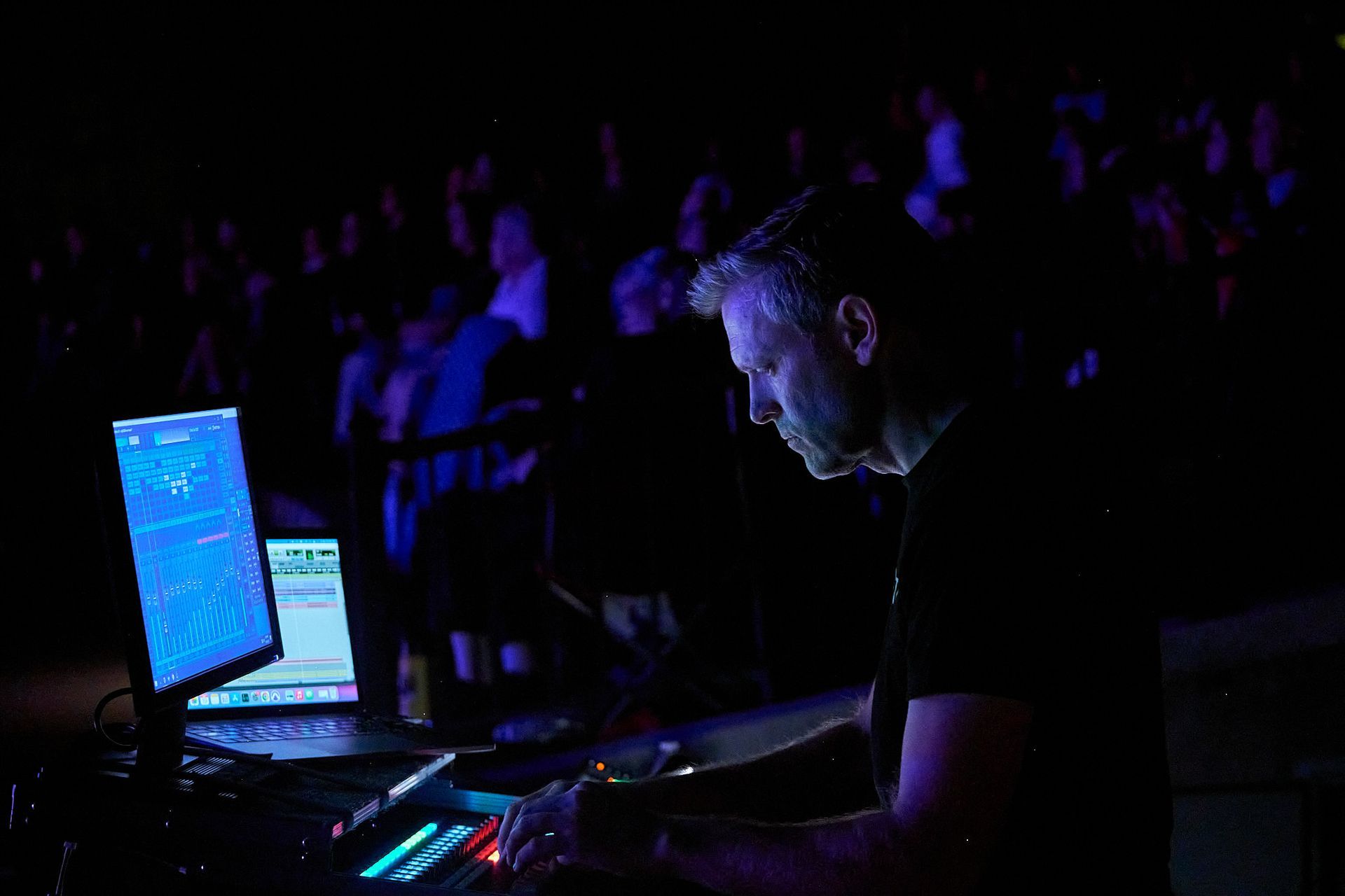 A focused person works at a mixing console with glowing computer screens in a dark, crowded theater setting.