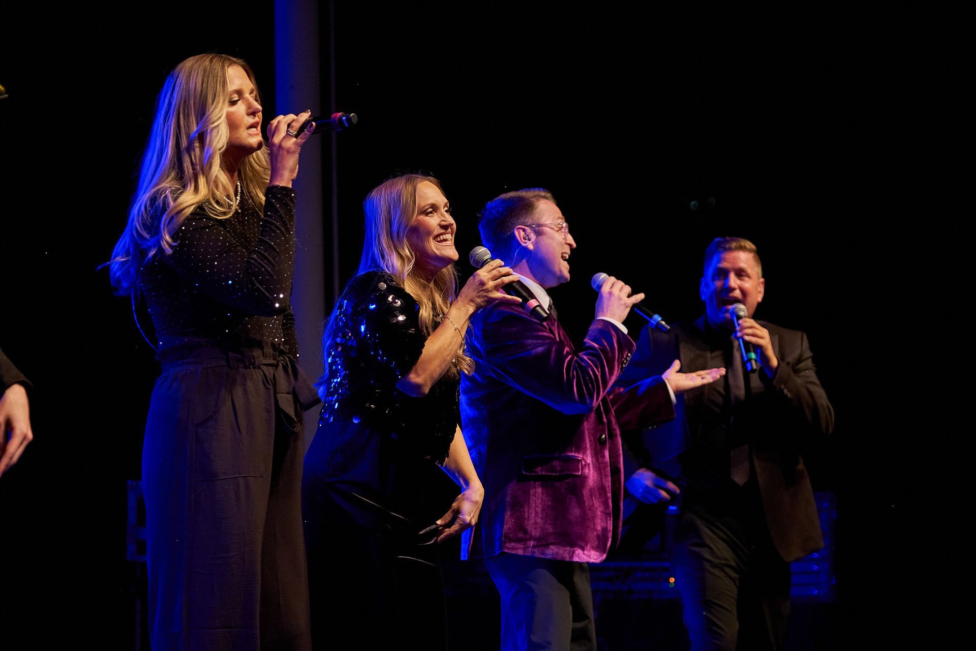 Four performers singing into microphones on a dark, dimly lit stage with blue accents.
