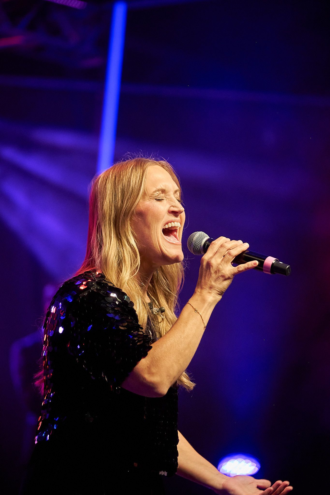 A singer with long blonde hair, wearing a black sequined top, singing into a microphone on a stage with blue lighting.