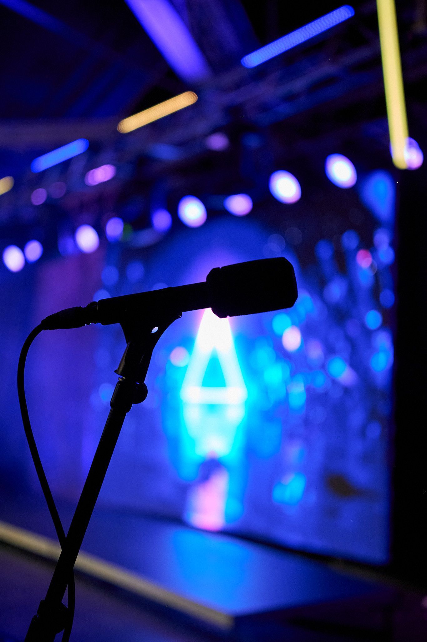 A silhouette of a microphone on a stand against a blurred, vibrant blue stage lit by glowing lights and a triangle icon.