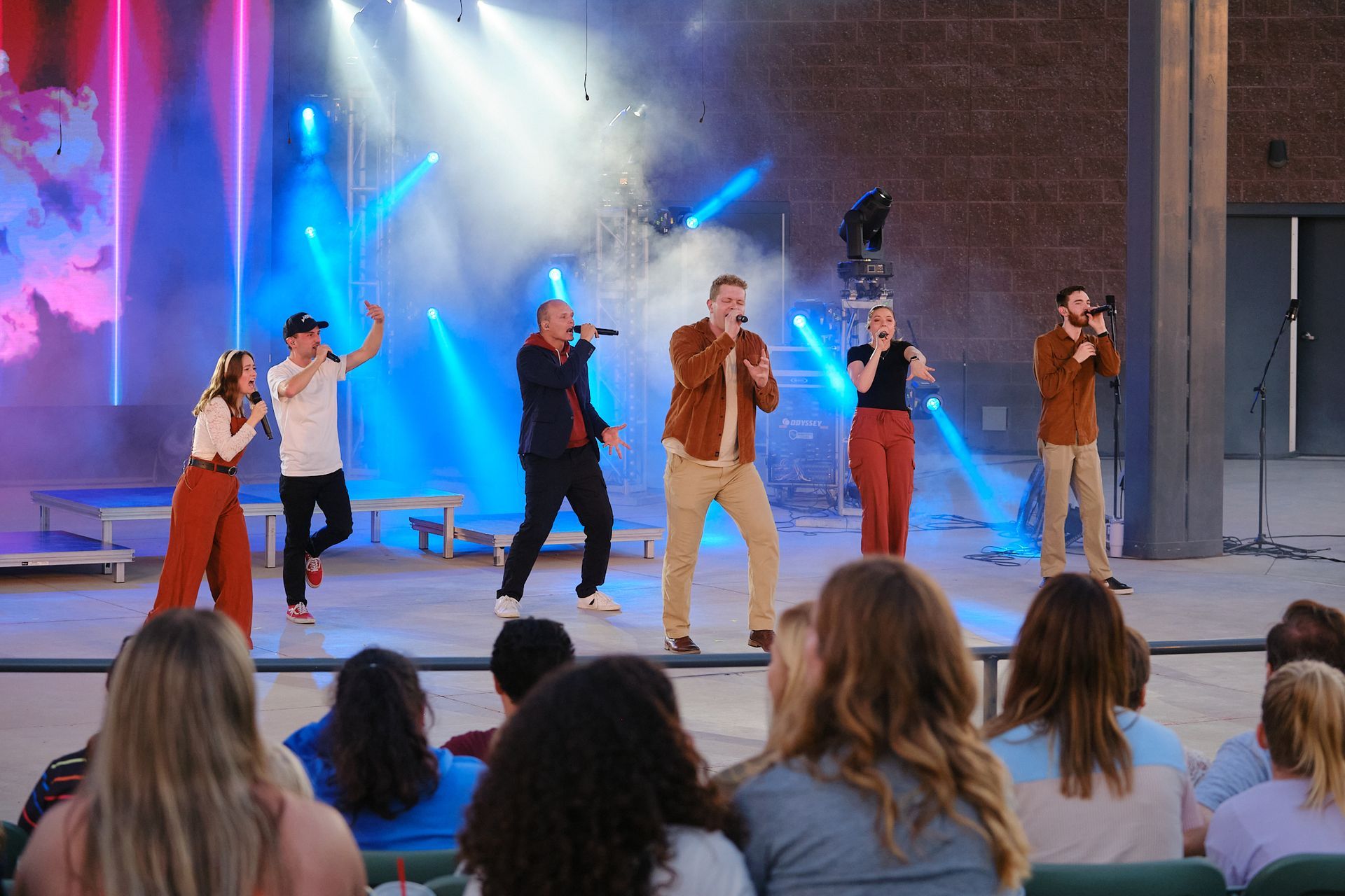 Five singers perform on a stage with blue lighting and fog before an audience seated in an outdoor venue.