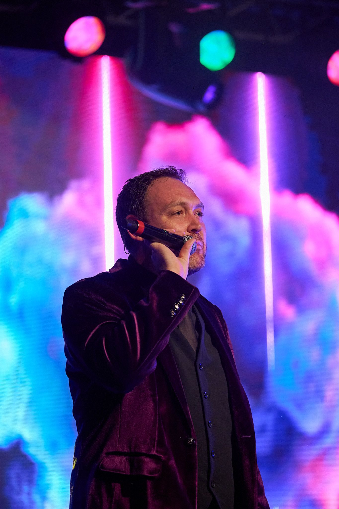 A person in a purple velvet jacket speaking into a microphone on stage, set against vibrant blue and pink smoke lights.